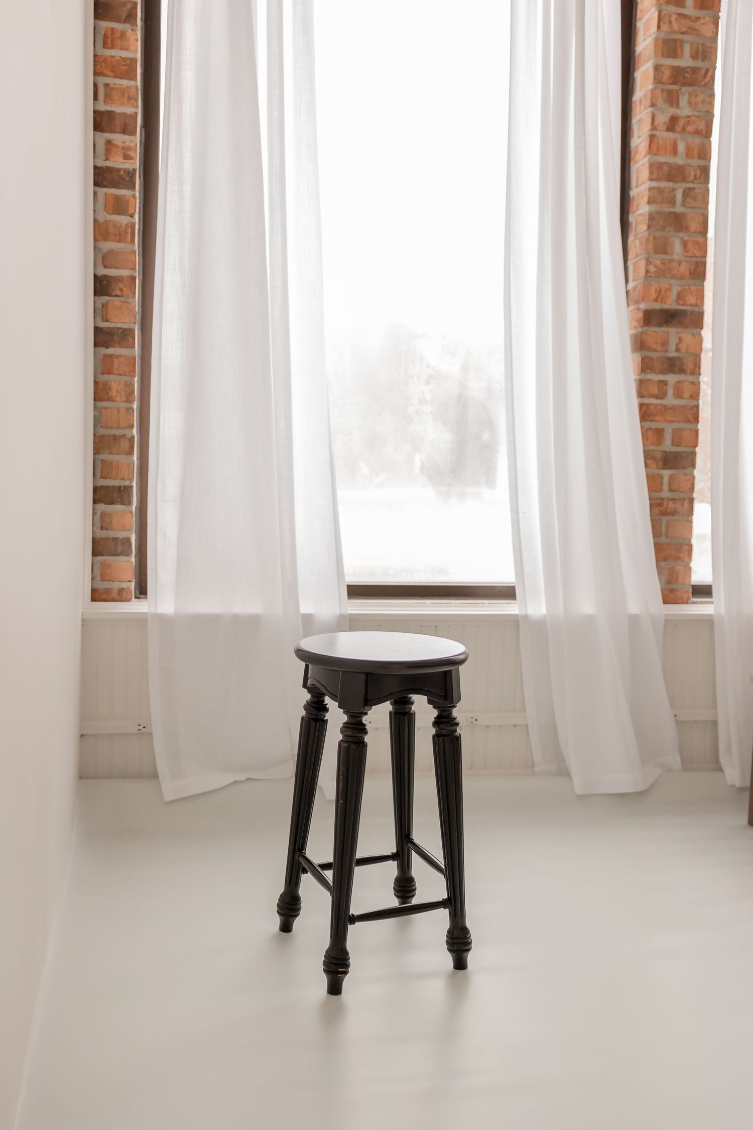Black wooden stool against exposed brick wall with white curtains and natural lighting in minimalist studio setting.