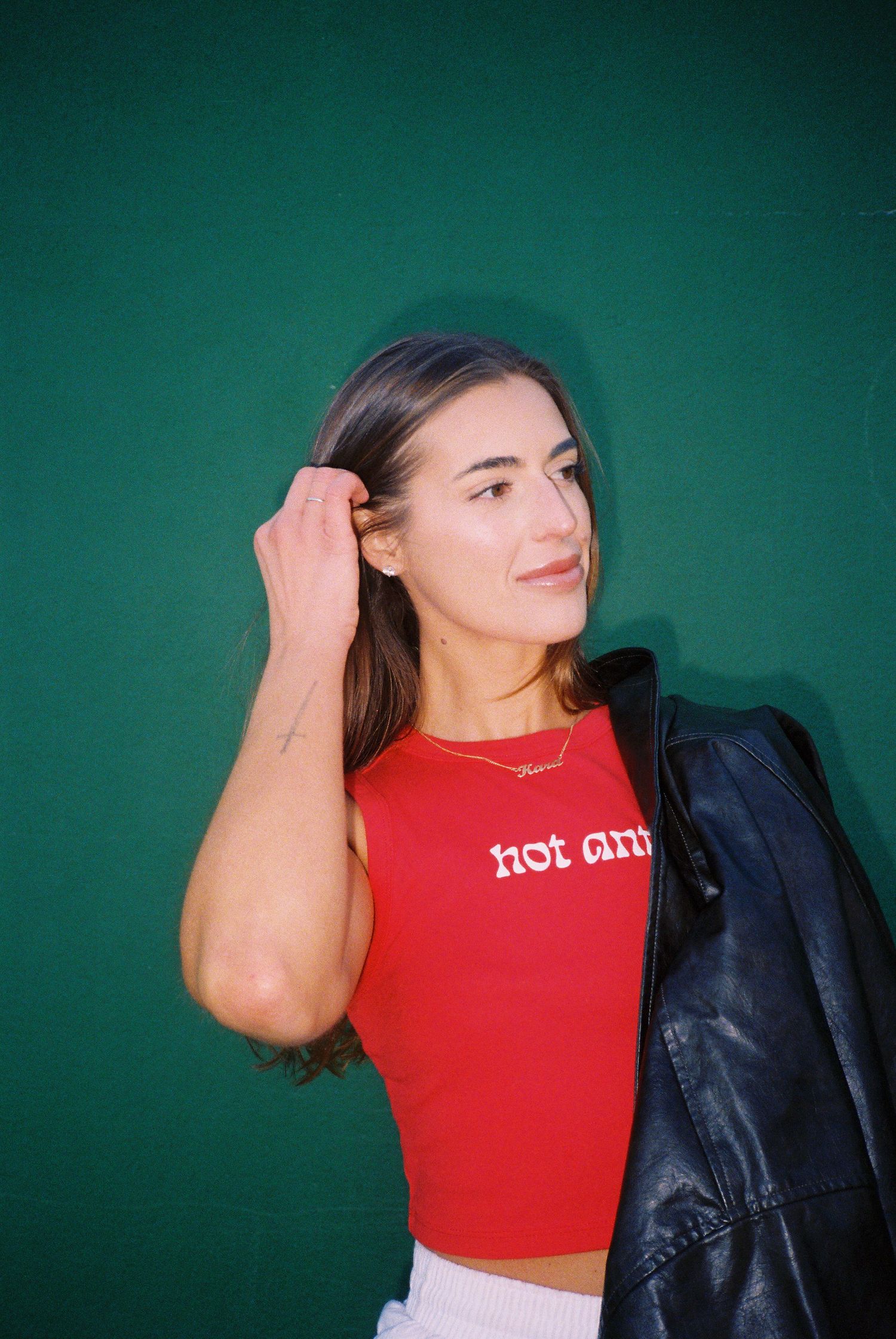 Young woman in red crop top and leather jacket posing against green wall.