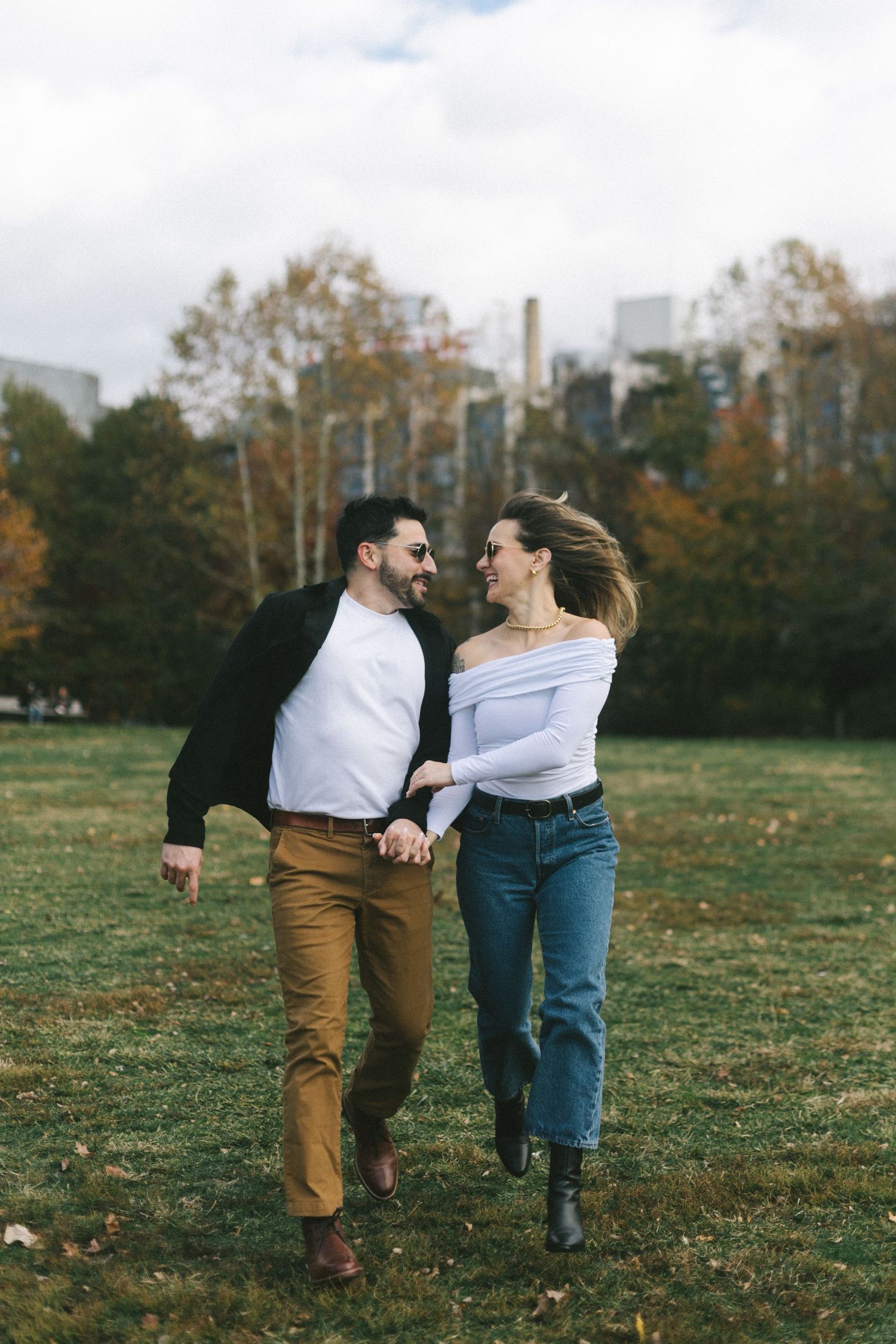 Happy couple holding hands and running through a green park with autumn trees and city buildings in the background.