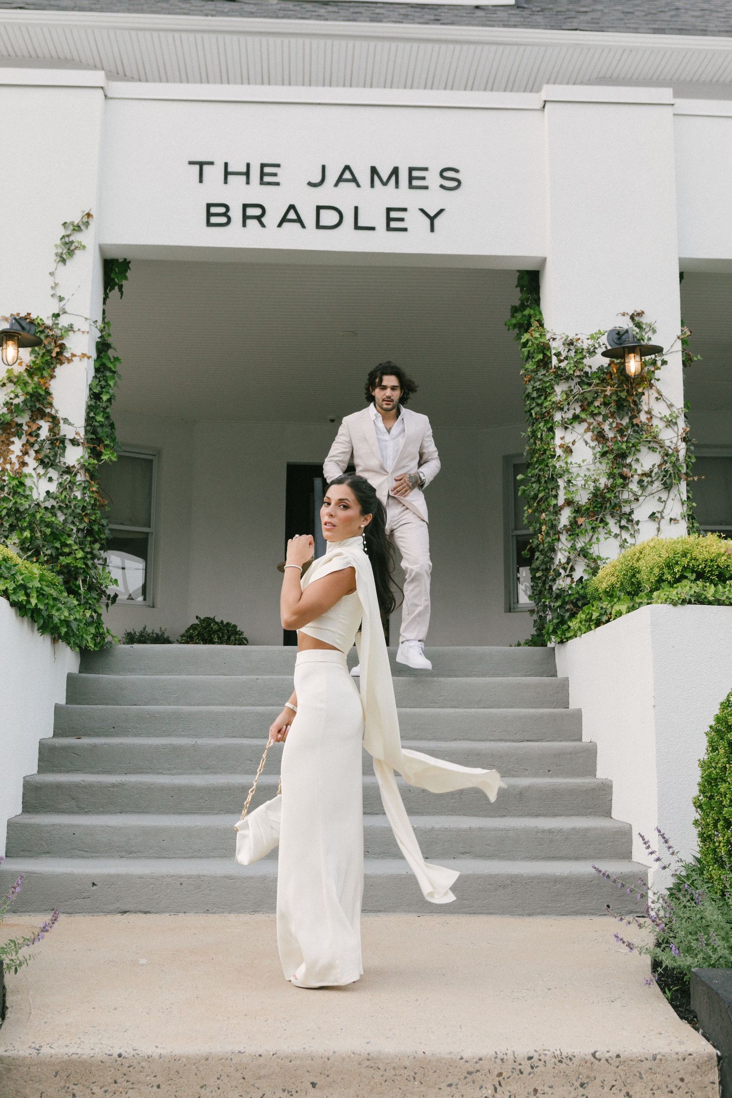 Couple in white outfits posing outside The James Bradley venue, woman in foreground with flowing sash, man on steps behind.