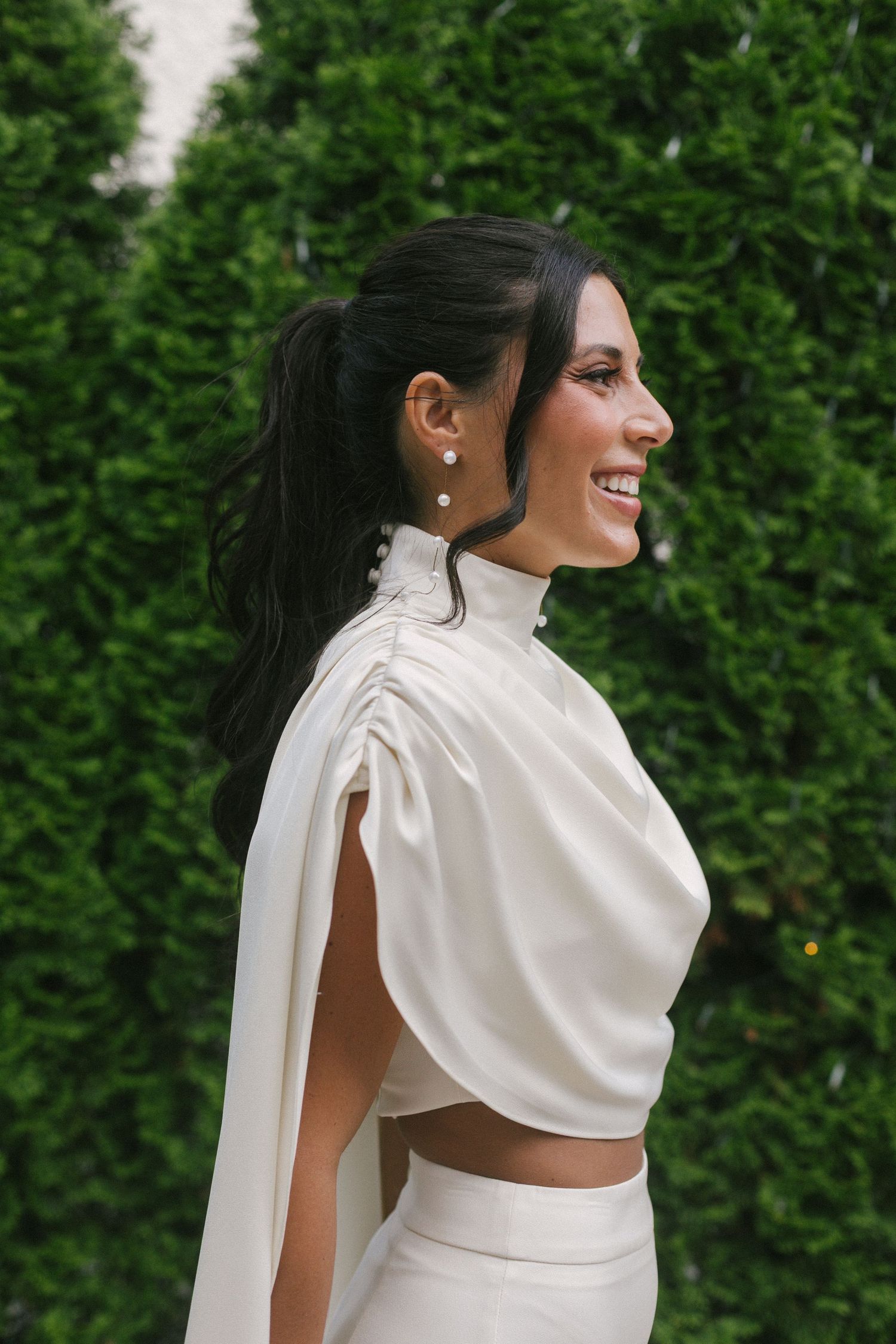 Smiling woman in white high-neck cape top and skirt, dark hair in ponytail, pearl earrings, posing outdoors by green hedges.