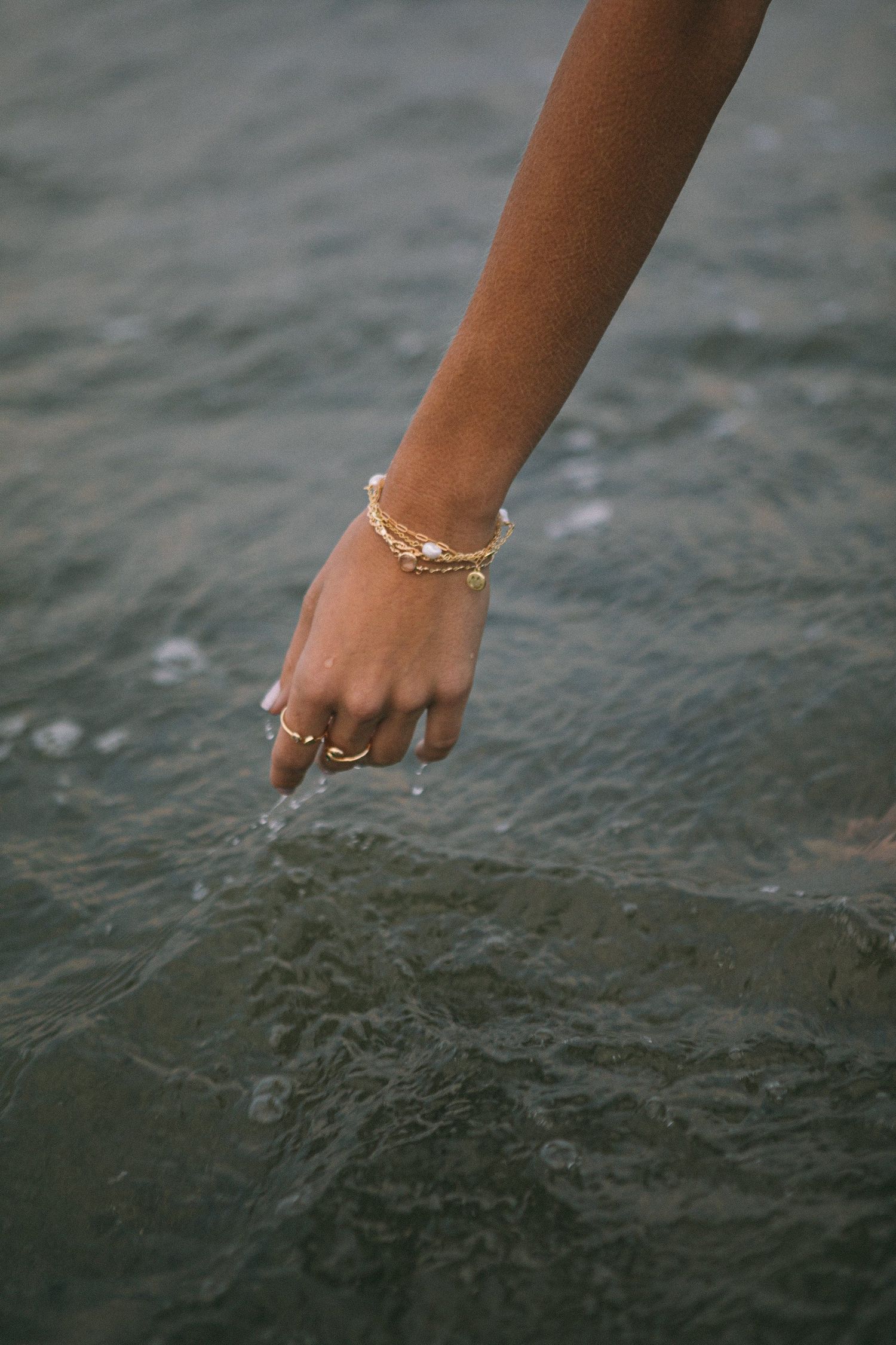 Hand with gold jewelry reaching down to touch water surface creating ripples.