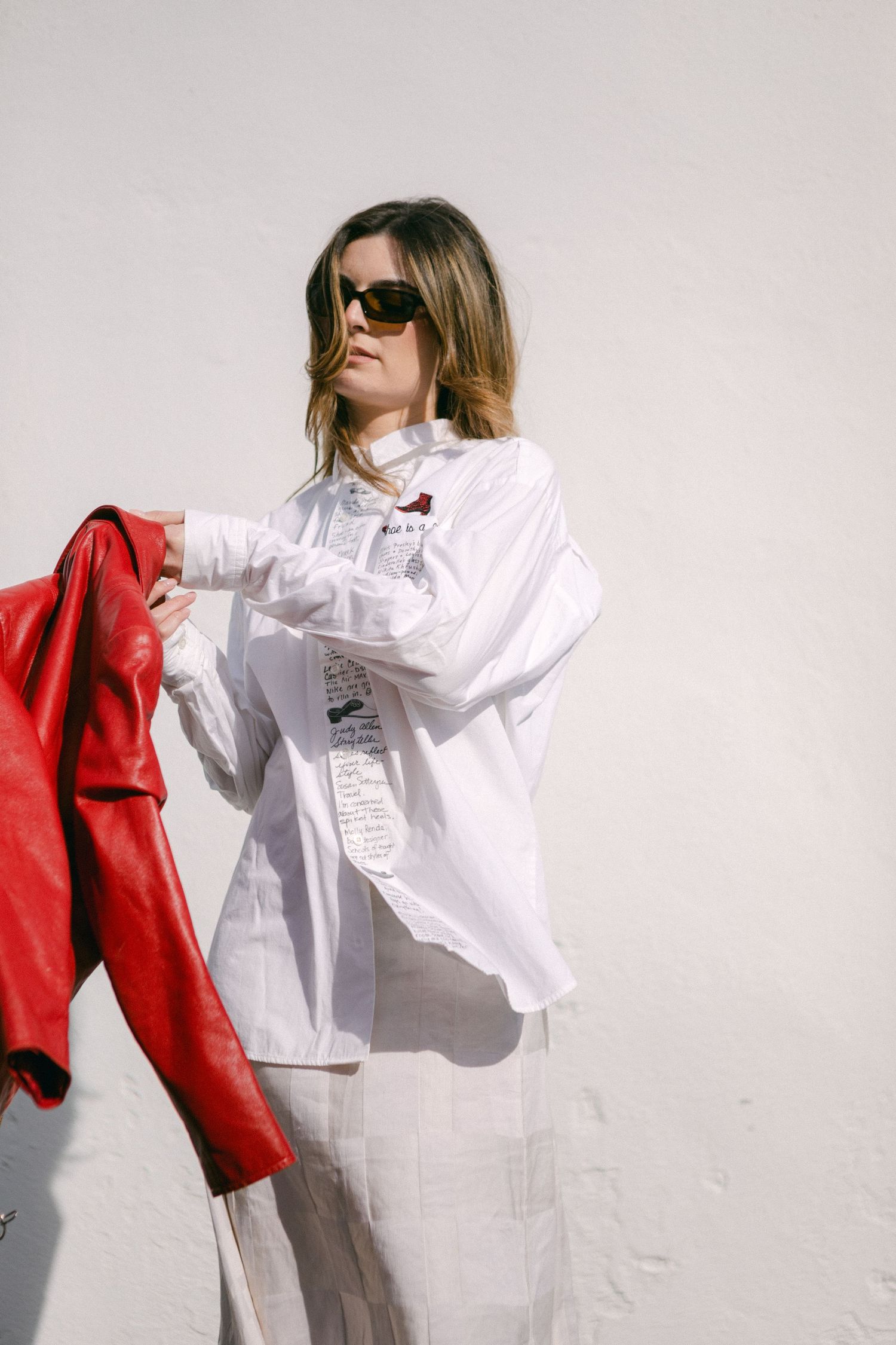 Fashion model wearing white shirt and red leather jacket poses dynamically against white wall in editorial style shoot.