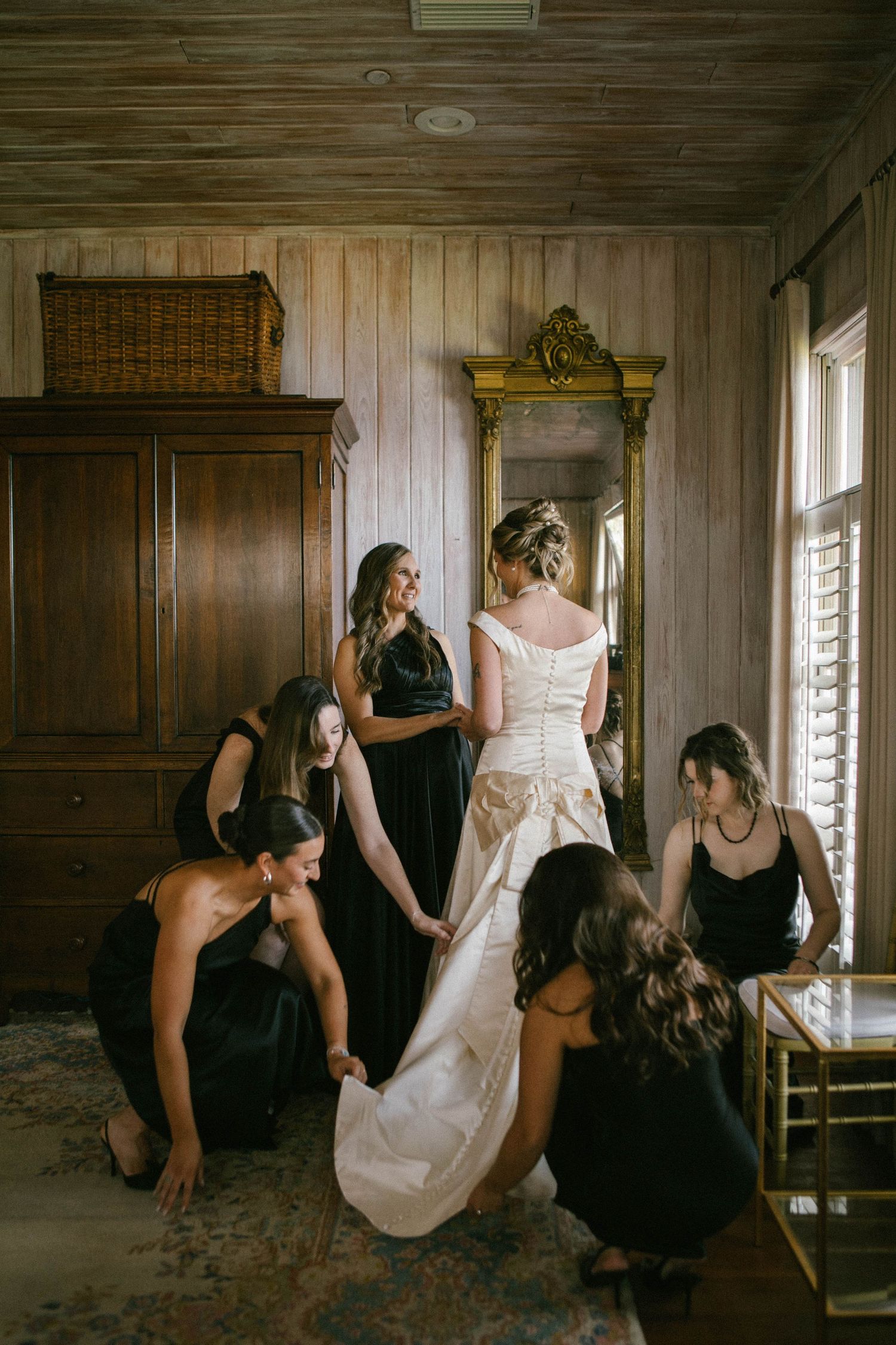 Bridesmaids in black dresses help prepare a white wedding gown in a vintage room.