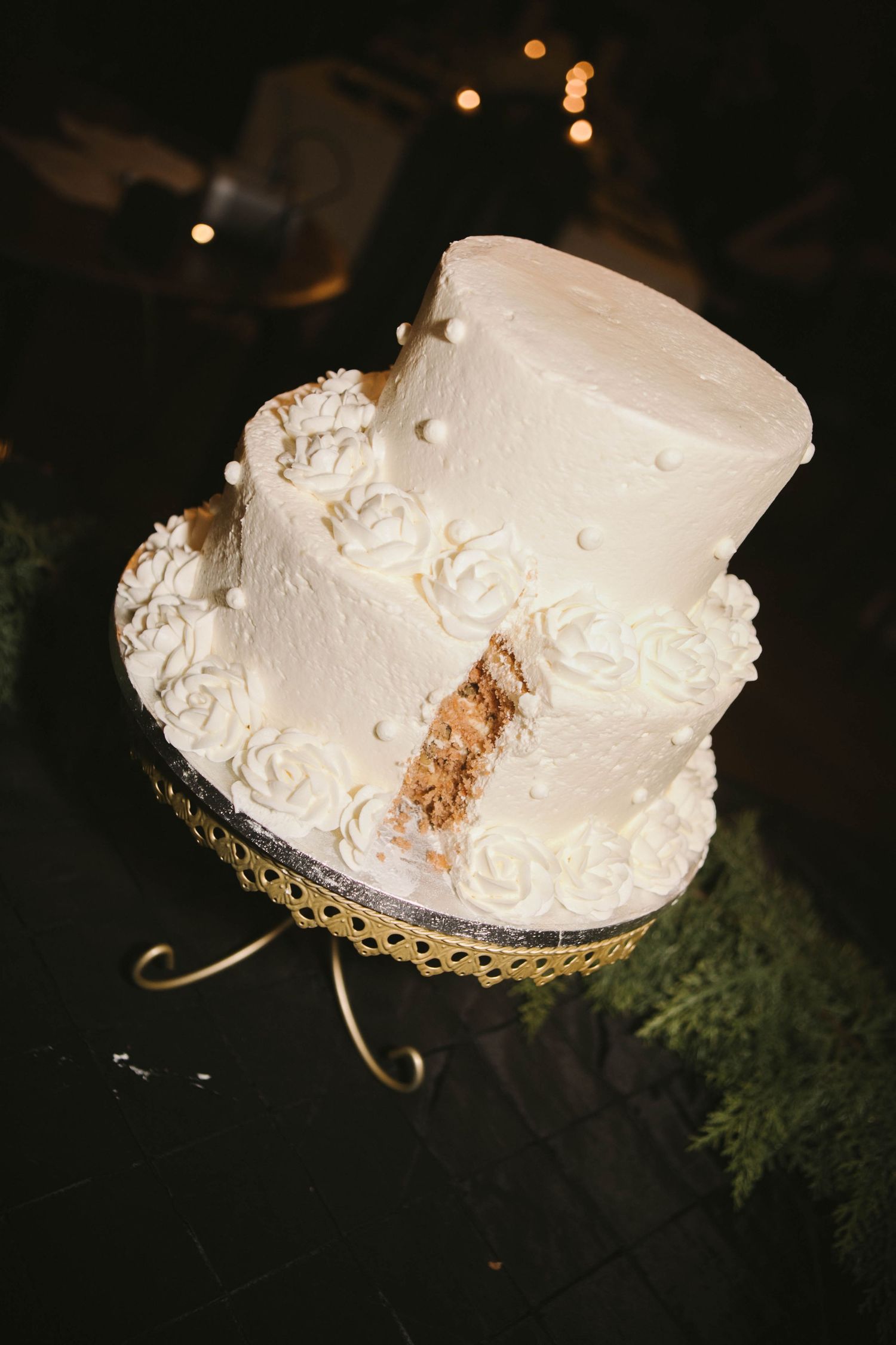 White wedding cake with floral decorations and pearls displayed on a gold stand against dark background.