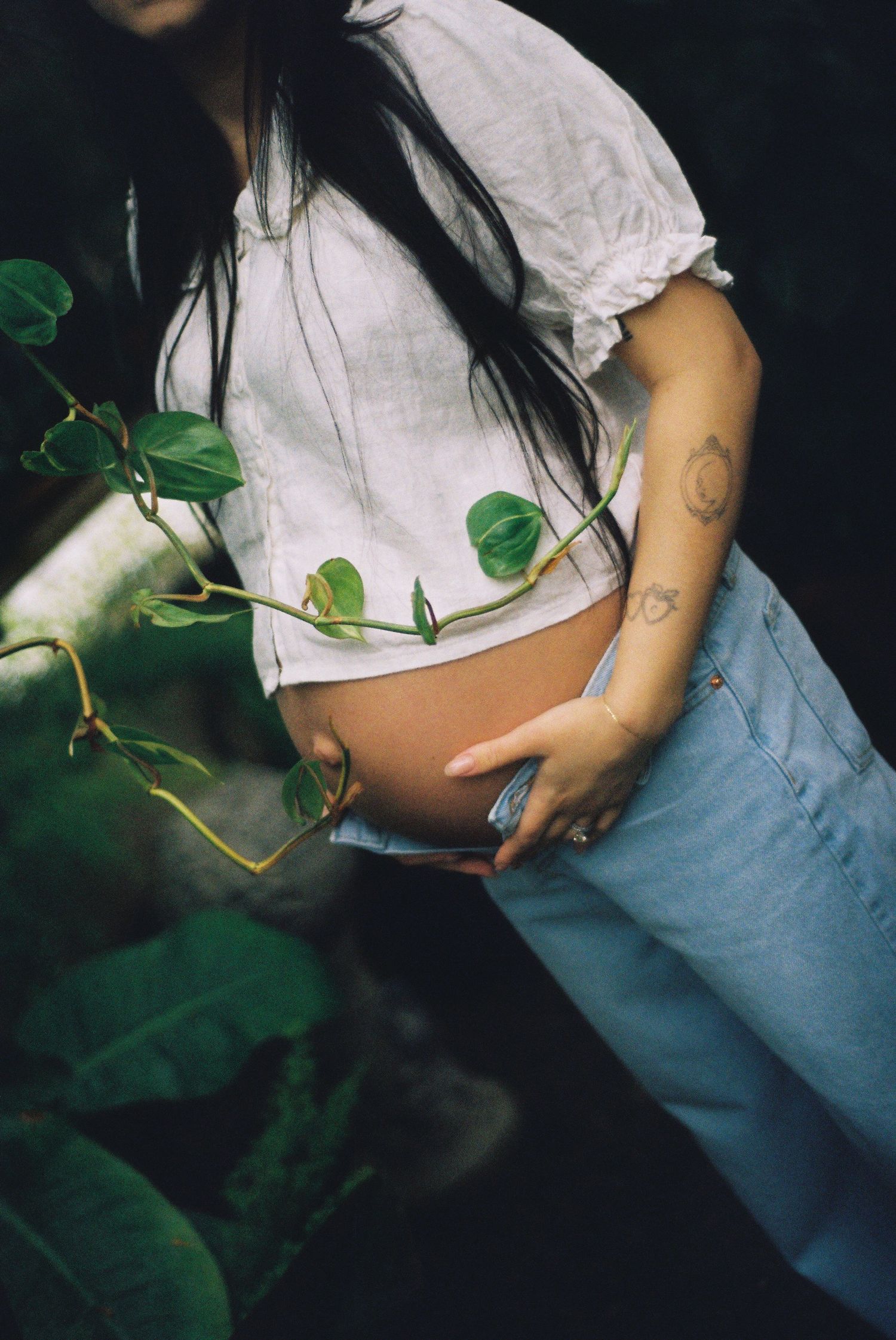 Pregnant woman in white crop top and jeans cradling baby bump surrounded by lush green tropical vines.