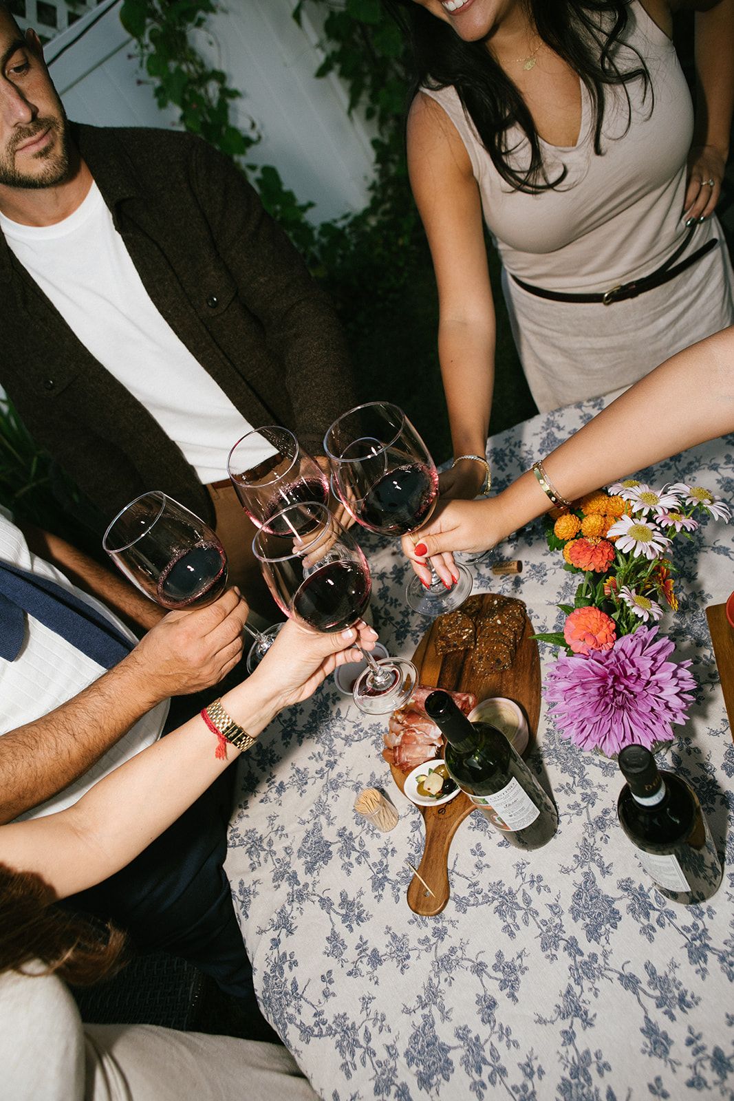 People gather around a floral tablecloth with wine glasses and flowers, creating an intimate dining atmosphere.