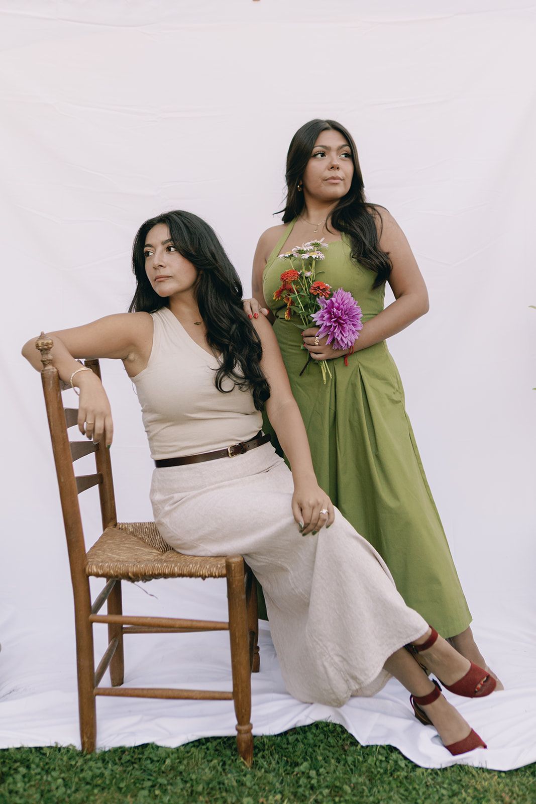 Two women in contrasting white and green dresses pose artistically on wooden chairs against a plain backdrop.