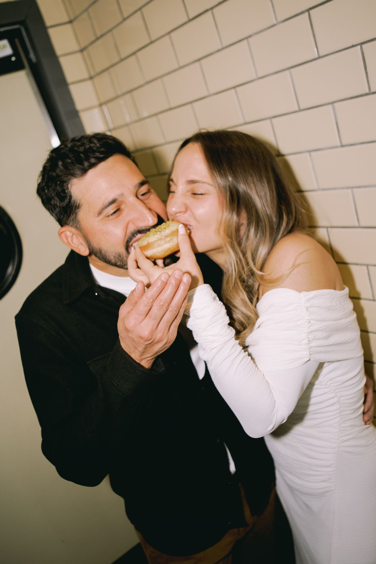 Couple sharing a donut kiss, bride in white off-shoulder dress and groom in black suit, romantic candid wedding moment.