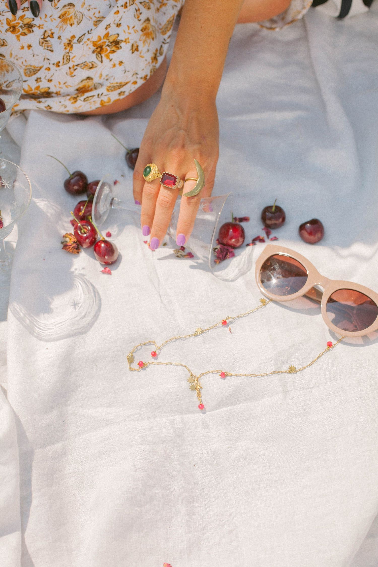 Hands arranging cherries and sunglasses on white linen tablecloth.