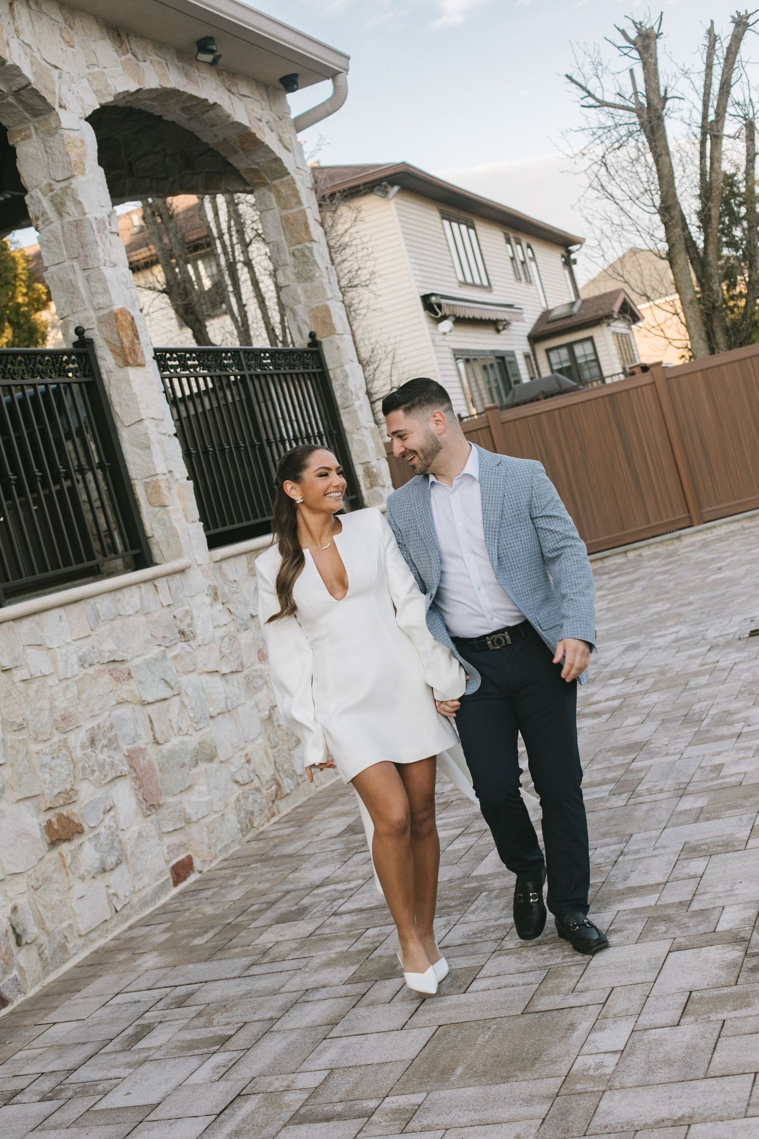 A smiling couple walks hand-in-hand on a stone patio, woman in white mini dress, man in gray blazer and dark pants.