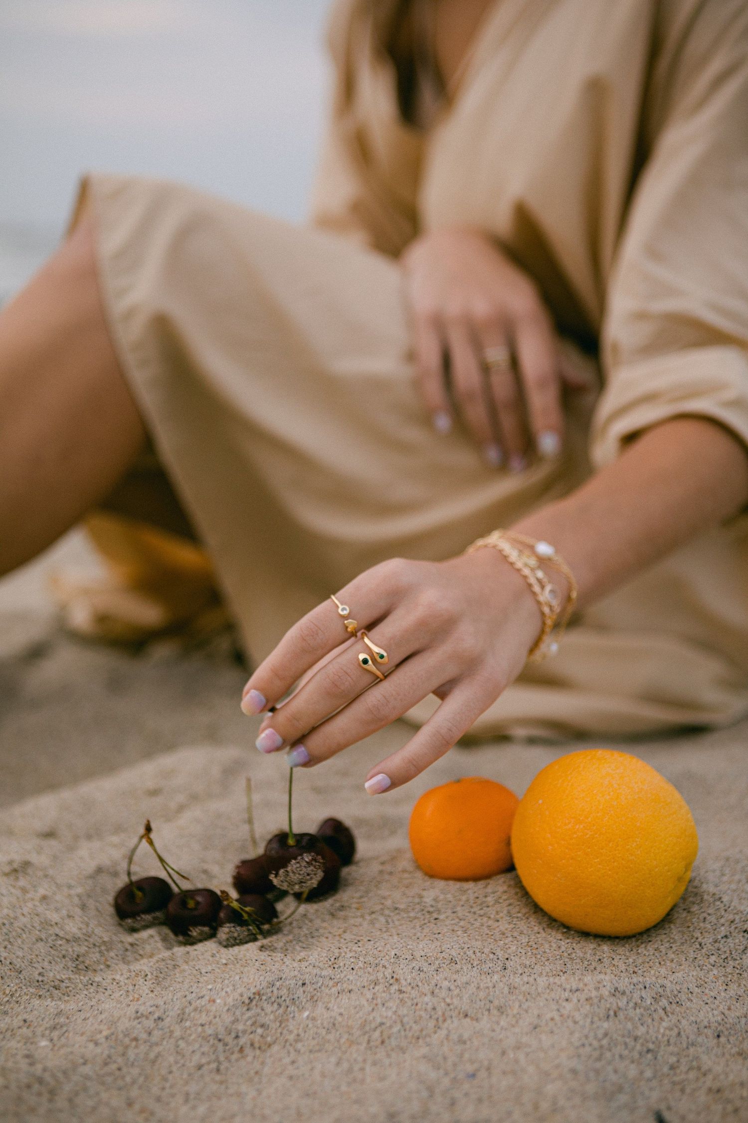 Hands with jewelry arranging oranges and dark objects on sandy surface.