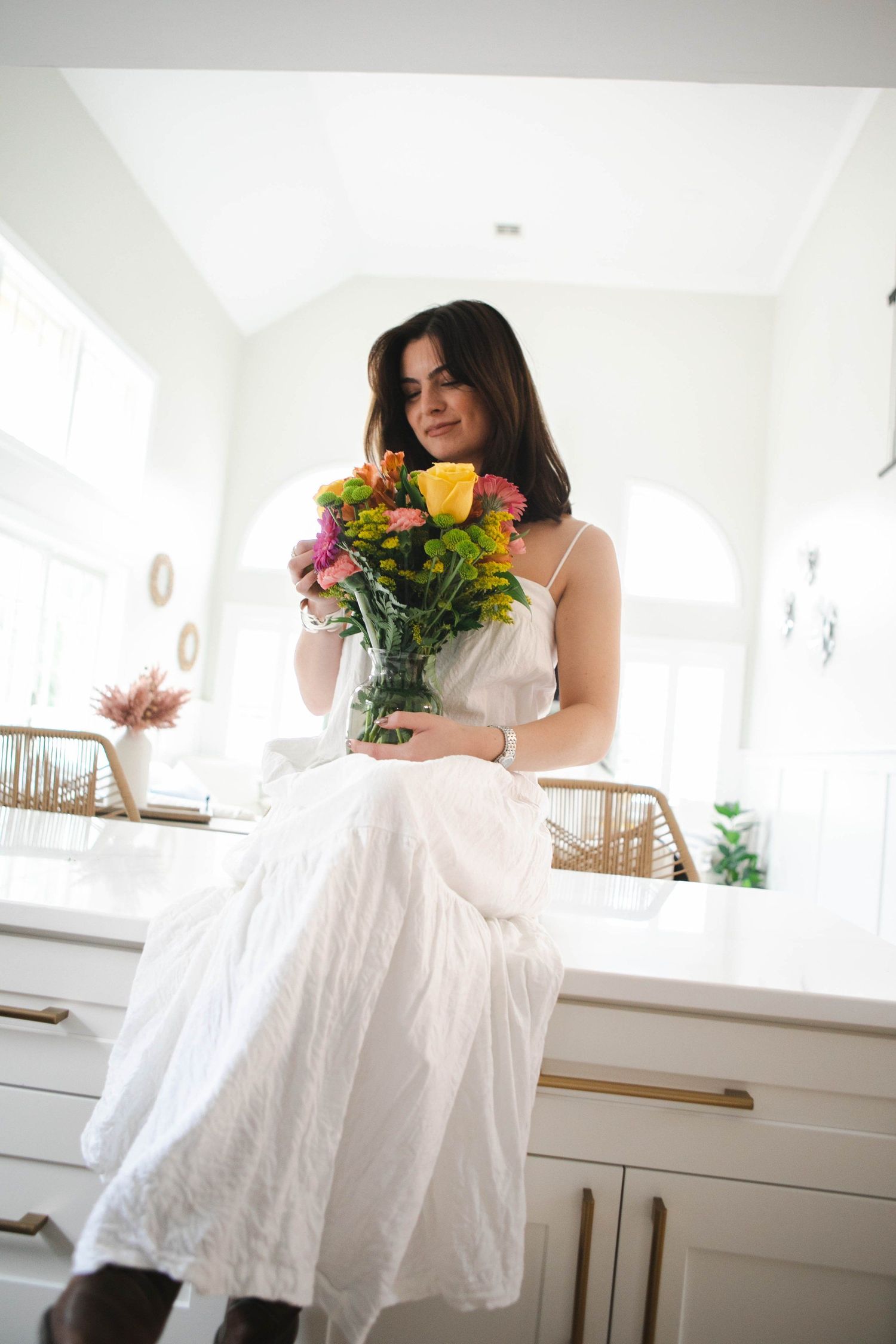 Woman in white dress holding colorful flowers in bright modern kitchen setting.