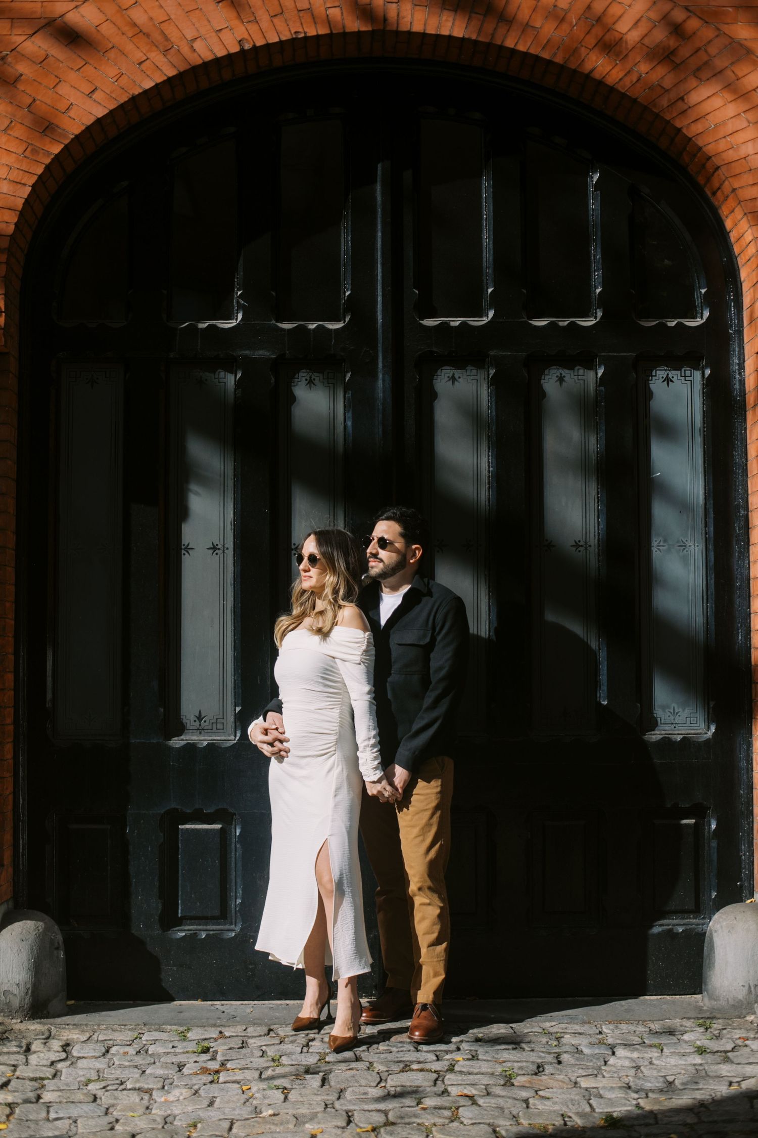 Couple holding hands in front of a large black arched door on cobblestones, woman in white dress, man in dark jacket.