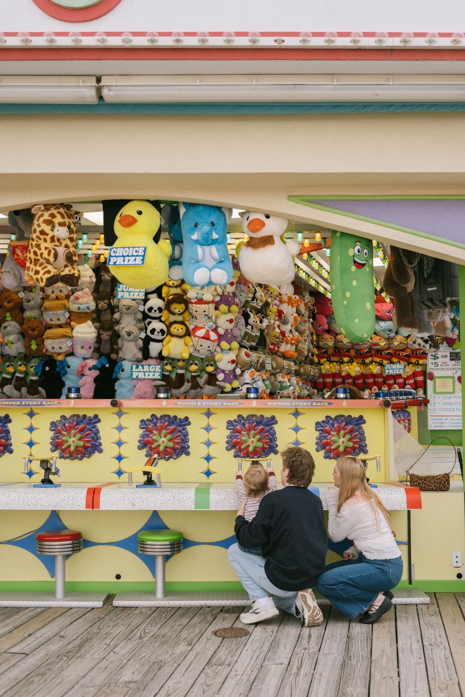 Family crouching together at a colorful carnival game booth filled with plush toy prizes on a wooden boardwalk.