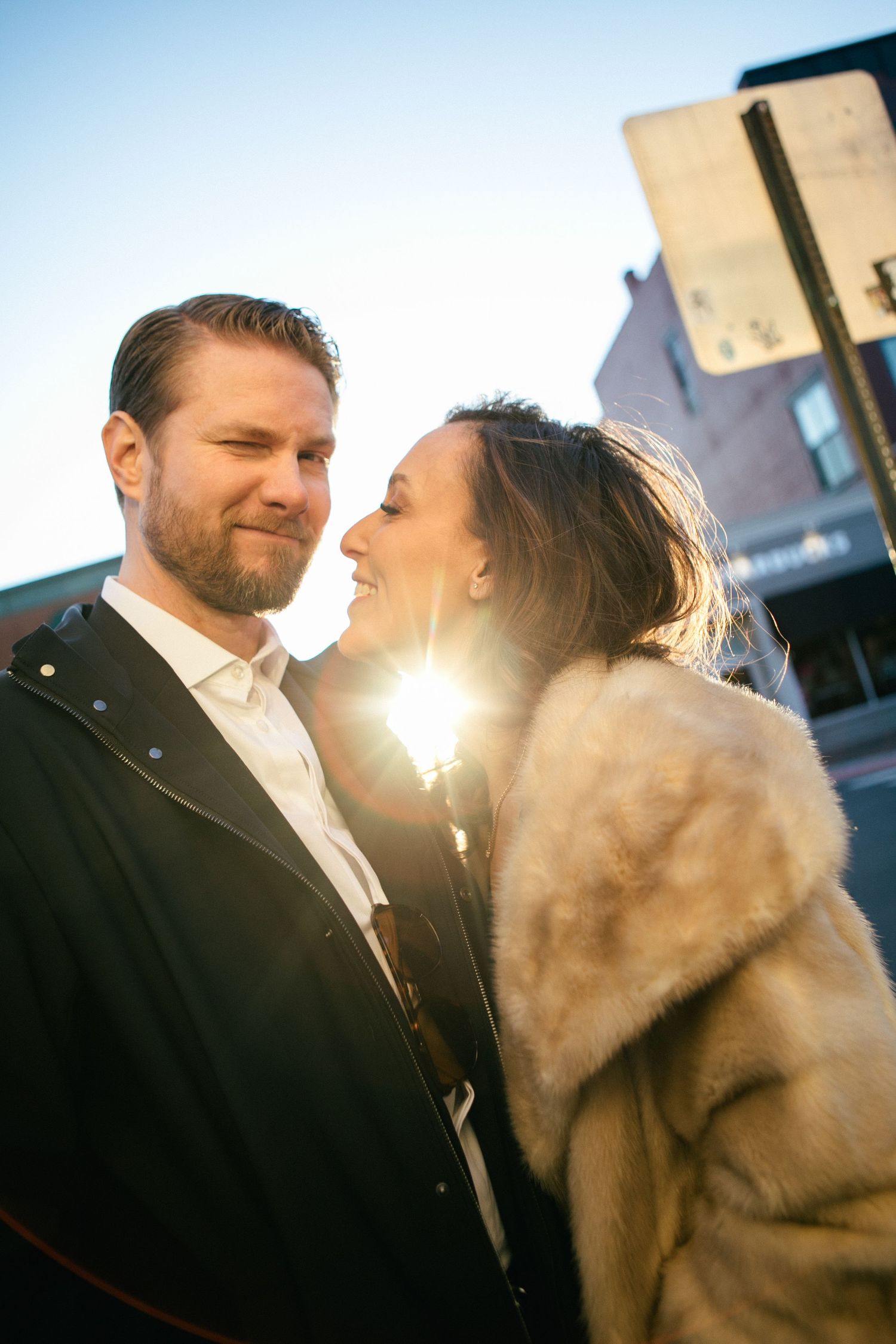 Couple sharing a romantic moment outdoors at sunset, woman in fur coat smiling at bearded man in black jacket.