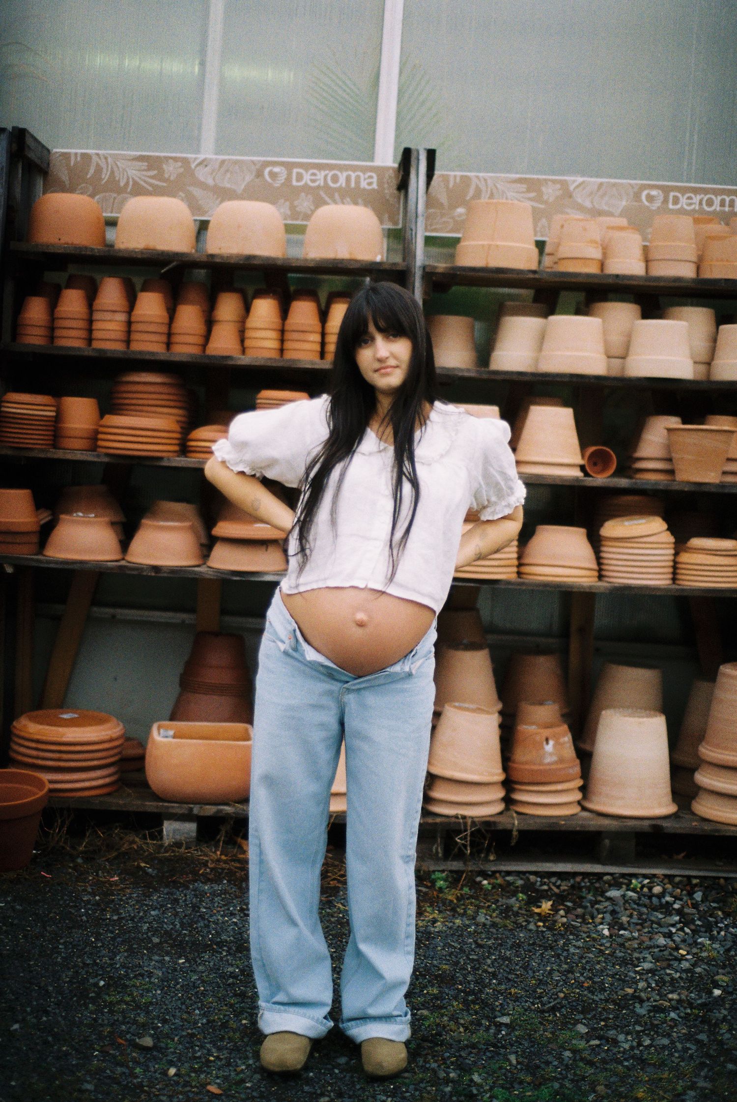 Pregnant woman in white top and light jeans poses in front of shelves of terracotta pots at a garden center.