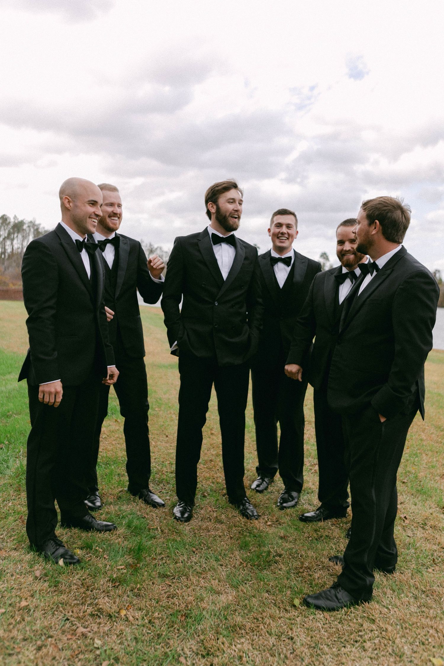 Groomsmen in black tuxedos gather on a grassy hillside.