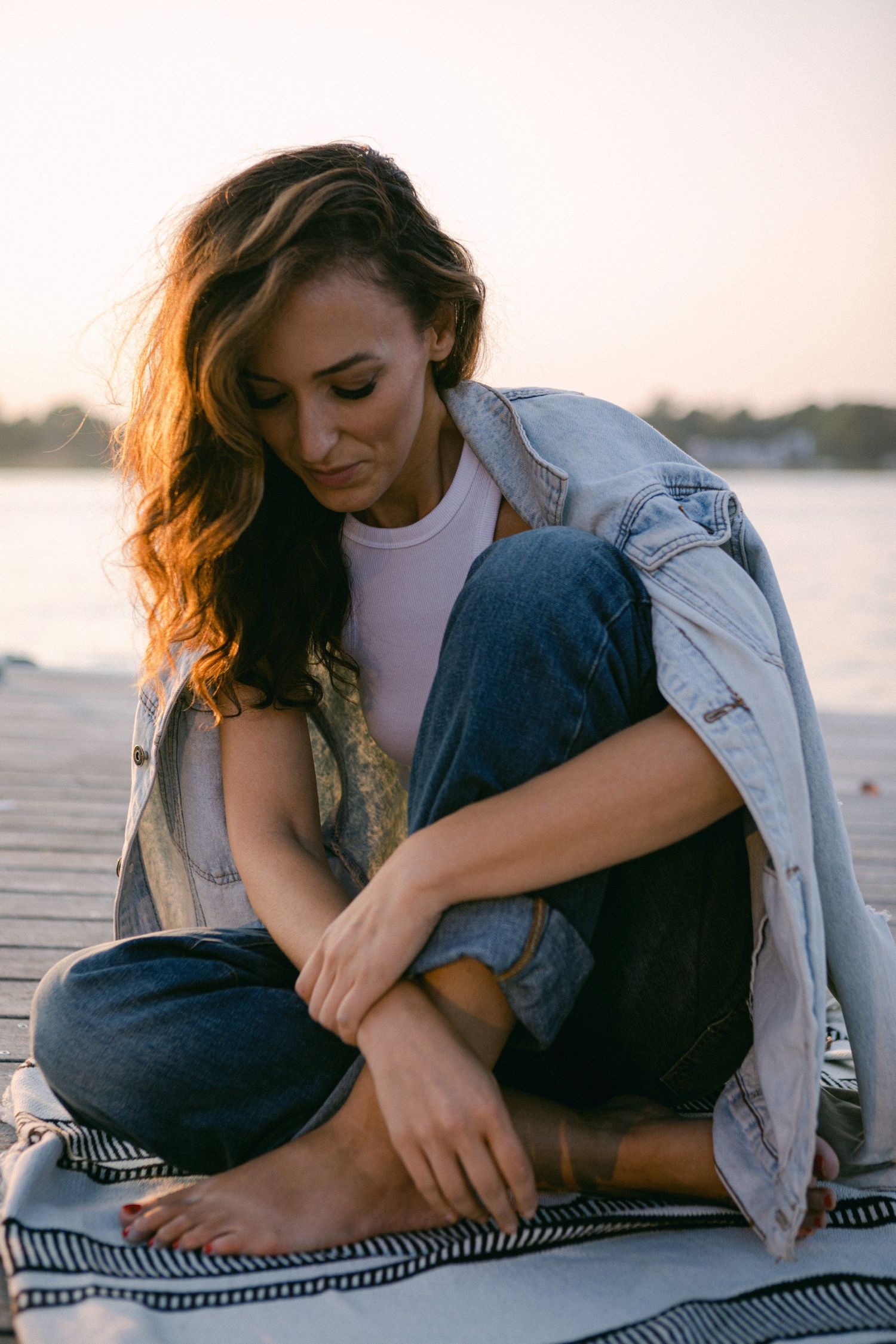 Person in denim jacket sitting on striped blanket by waterfront at sunset.