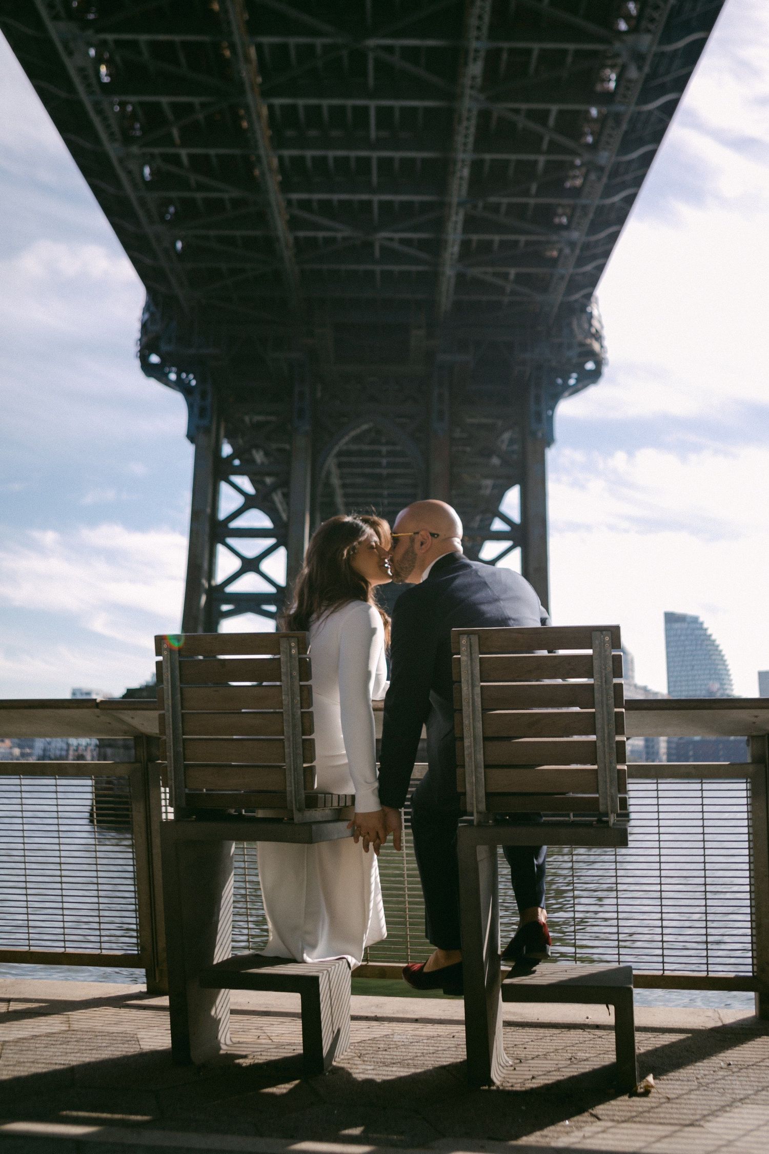 Couple sitting on wooden benches under a bridge with city skyline views.