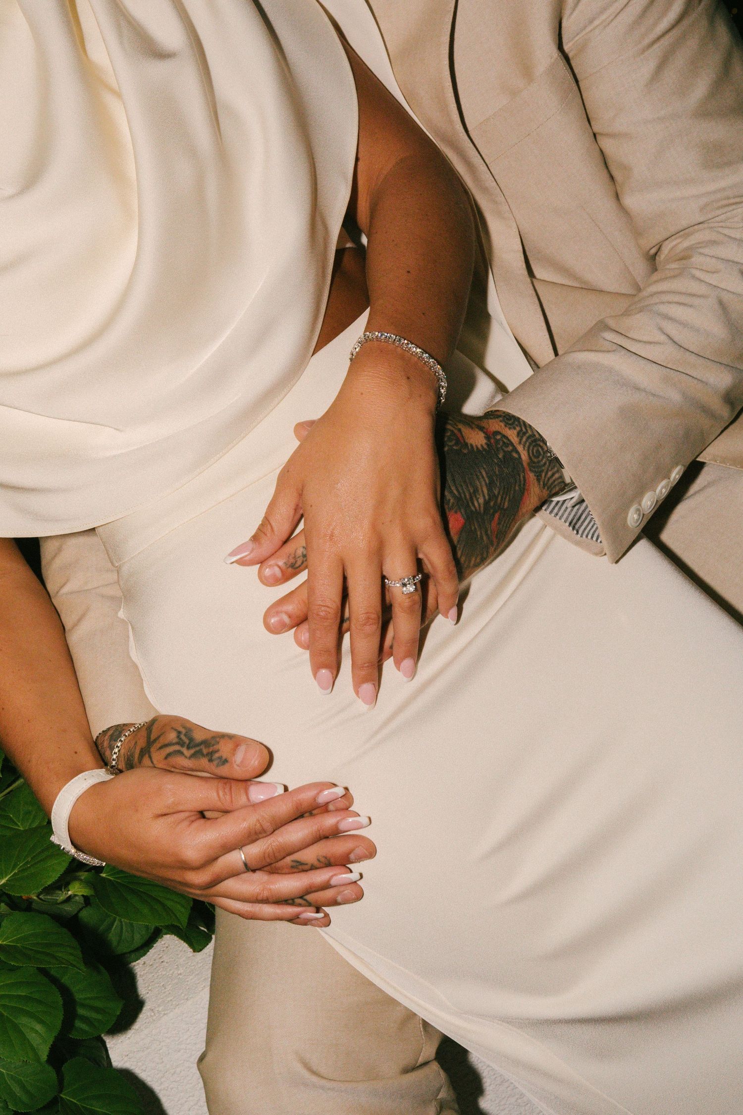 Close-up of couple's tattooed hands with diamond ring and bracelet, wearing beige suit and white wedding dress.