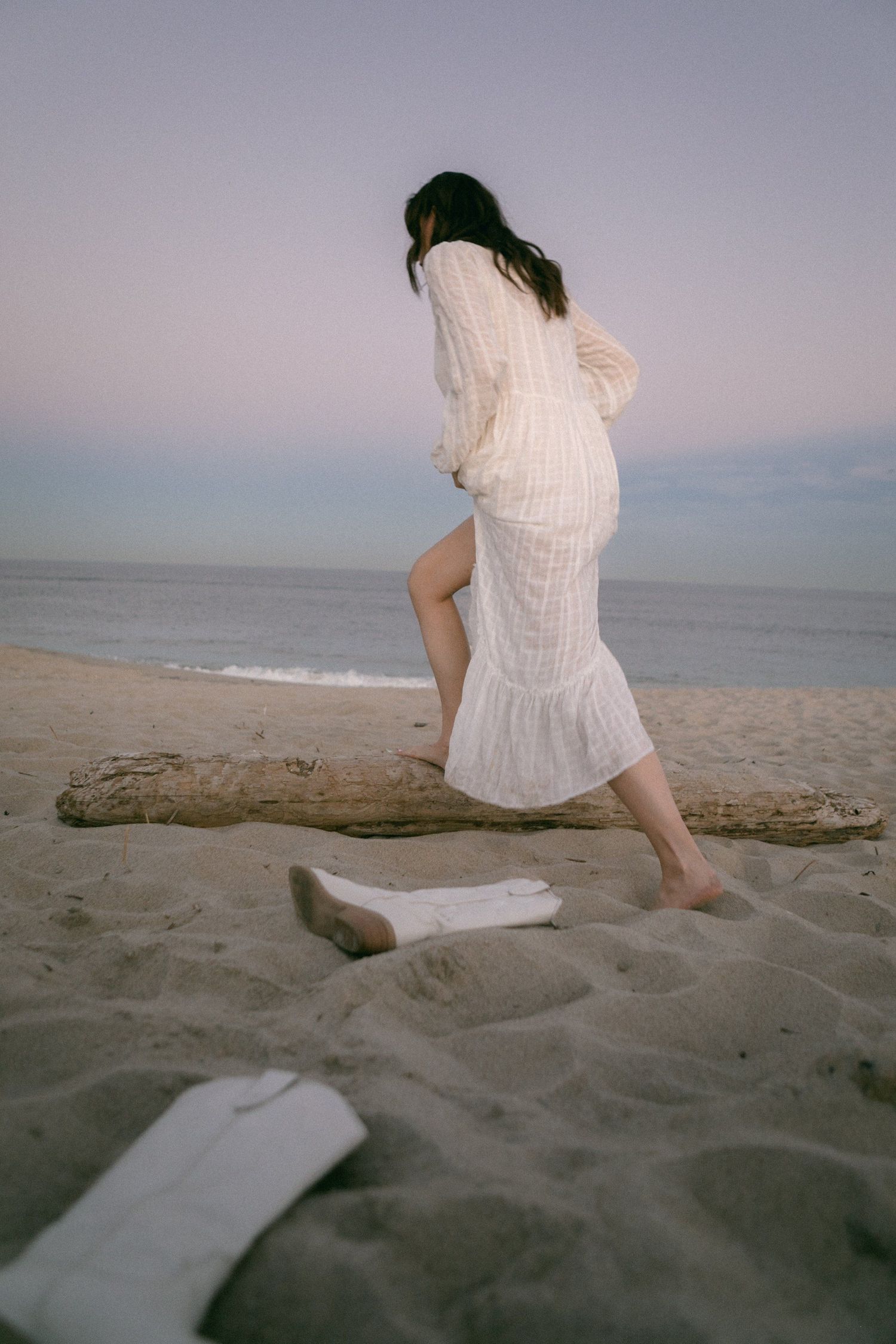 Person in white dress sitting peacefully on beach log at dusk.