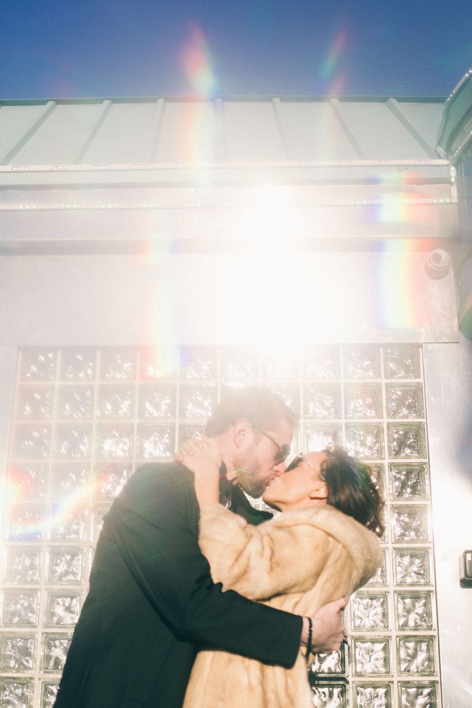 Couple sharing a romantic kiss outdoors with sunlight and rainbow lens flare, woman in fur coat, glass block wall backdrop.