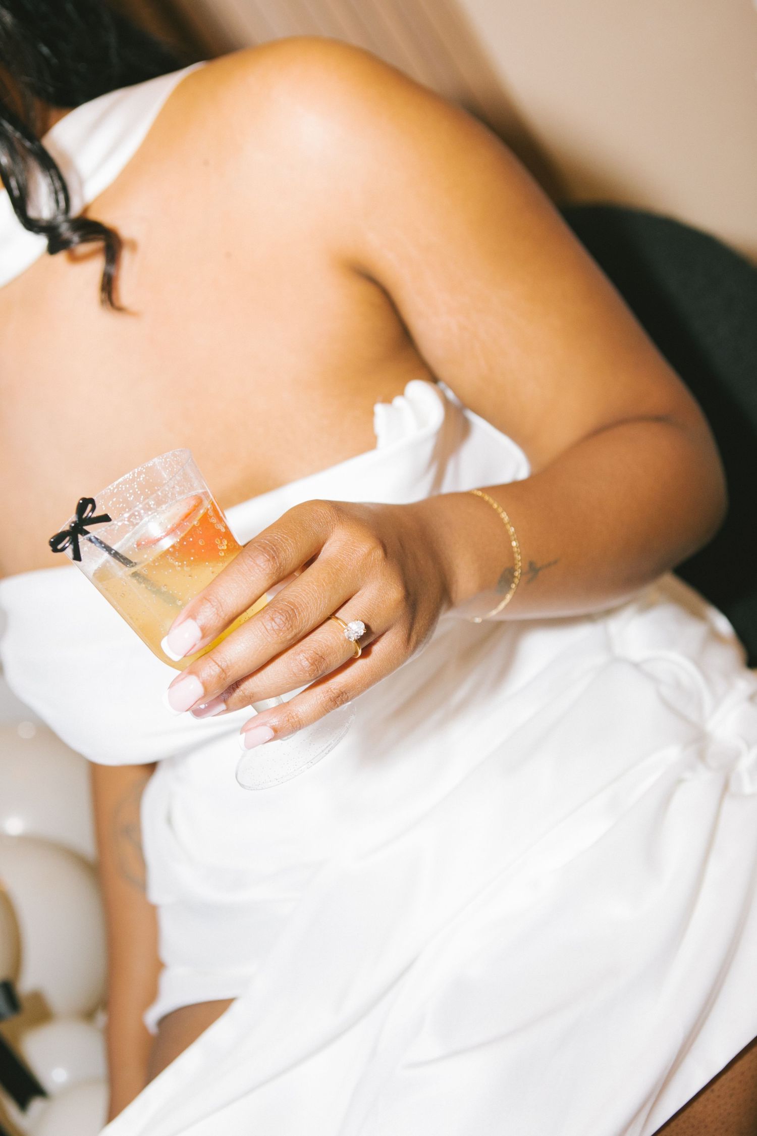 Bride in white dress holding a cocktail garnished with a bow, wearing a diamond ring and gold bracelet.