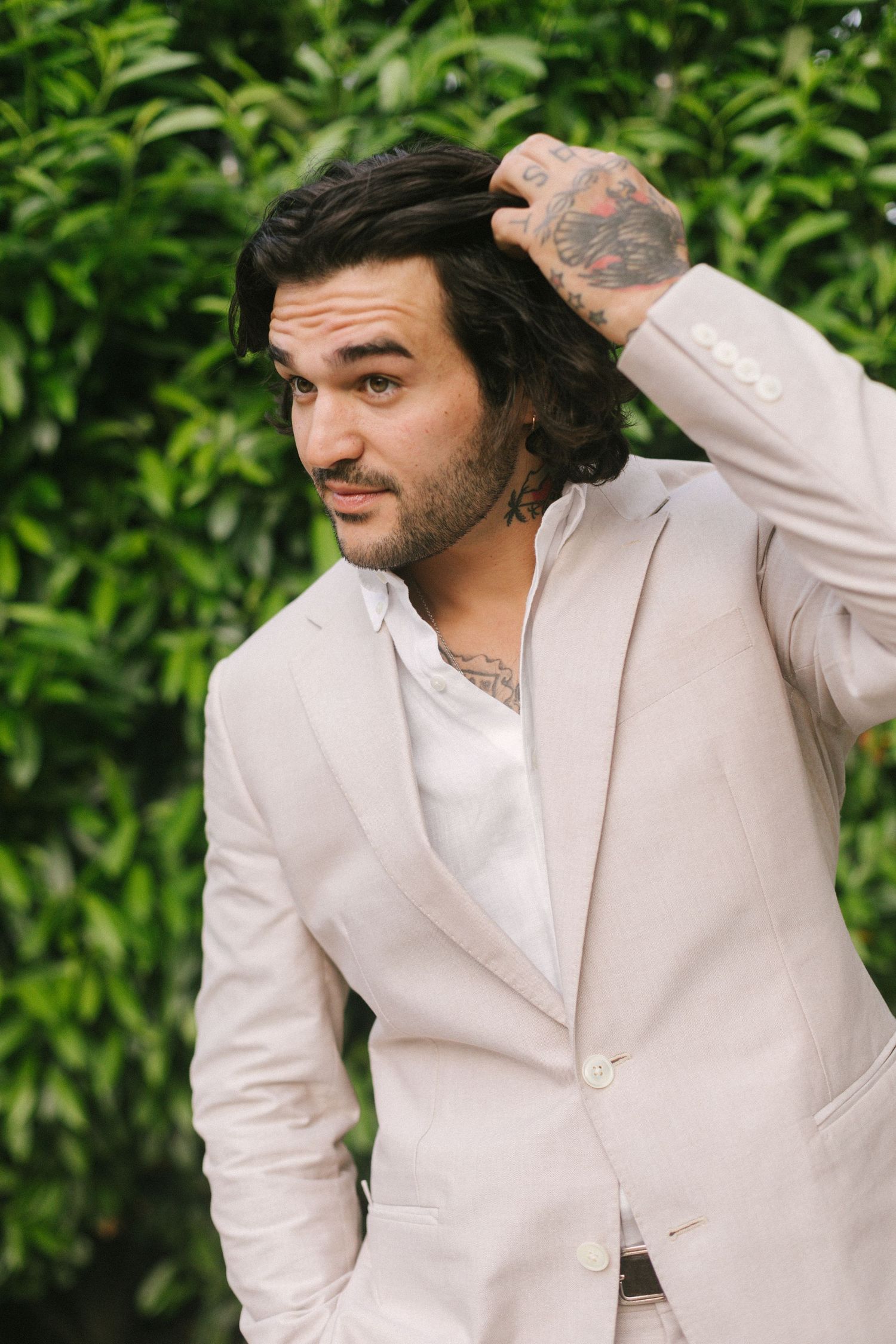 Tattooed man in beige linen suit running hand through dark hair against green leafy background.