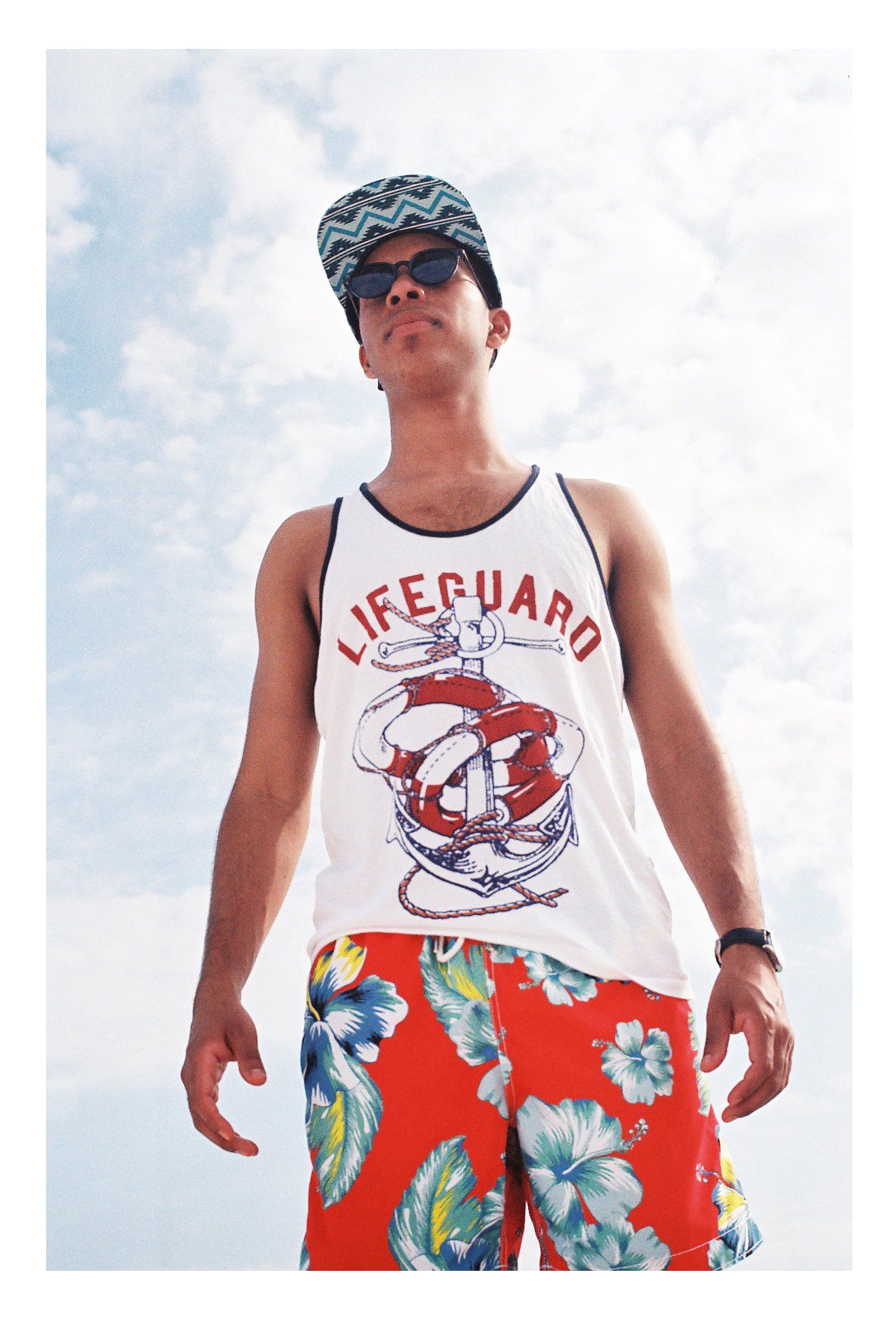 Person wearing a lifeguard tank top and floral swim trunks poses against blue sky.