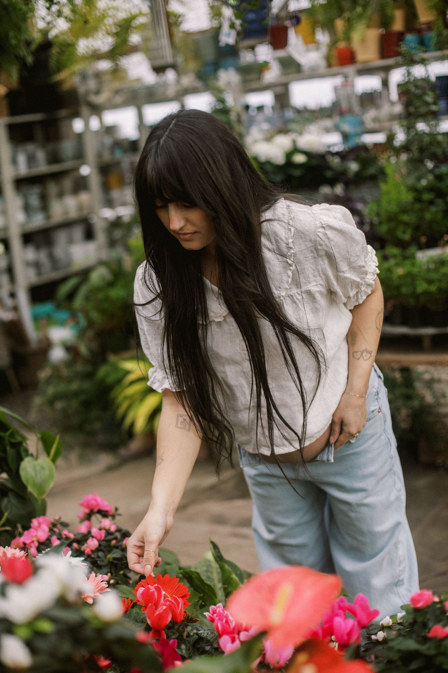 Dark-haired woman in white blouse and jeans bending over colorful flowers in a lush garden shop.