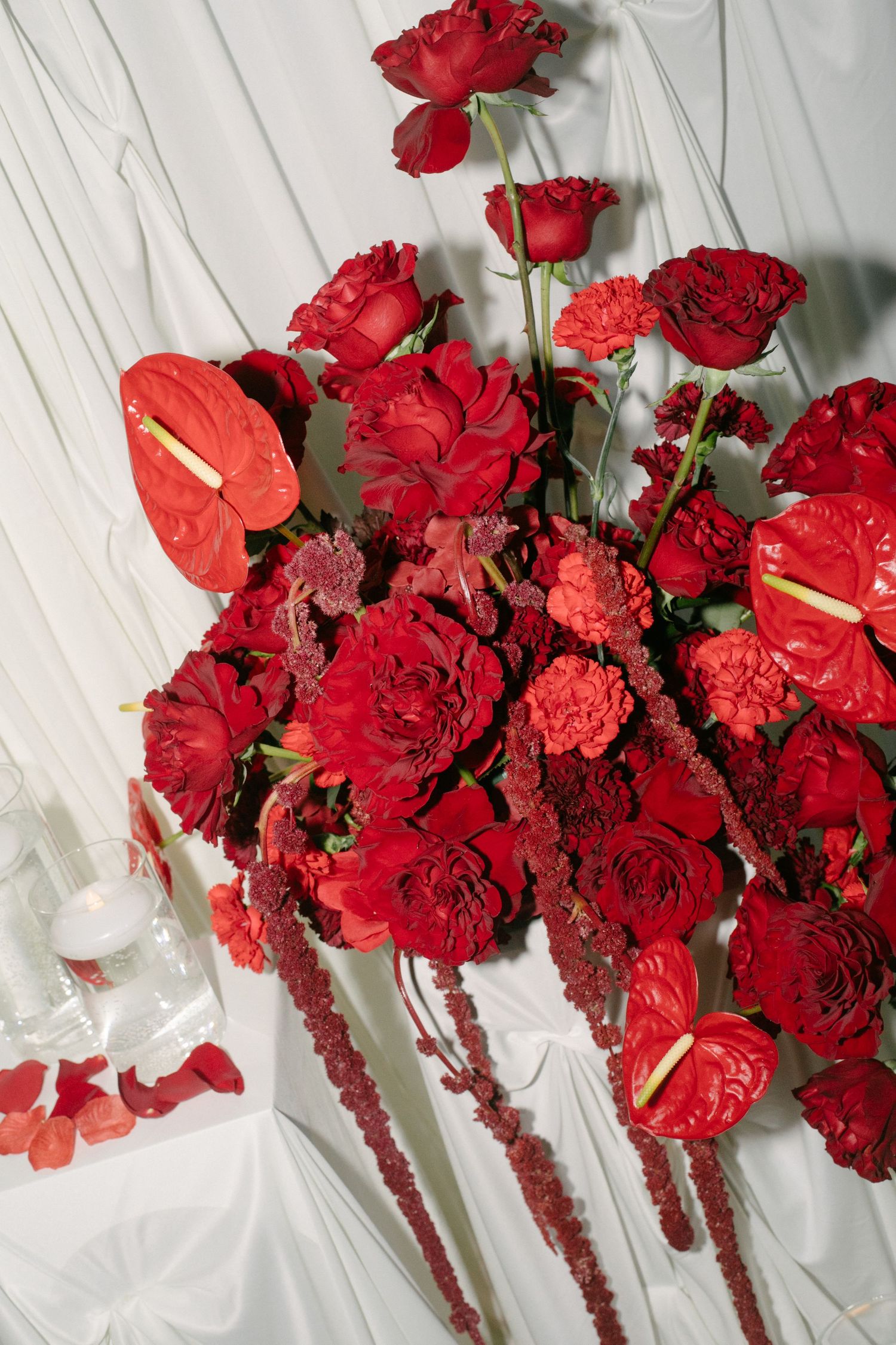 Lush arrangement of red roses, anthuriums, carnations, and amaranthus on white fabric backdrop.