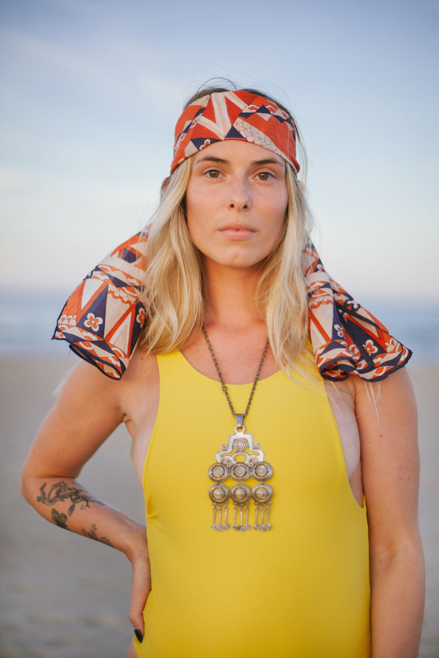 Person in yellow tank top wearing decorative headscarf and statement necklace against sky background.