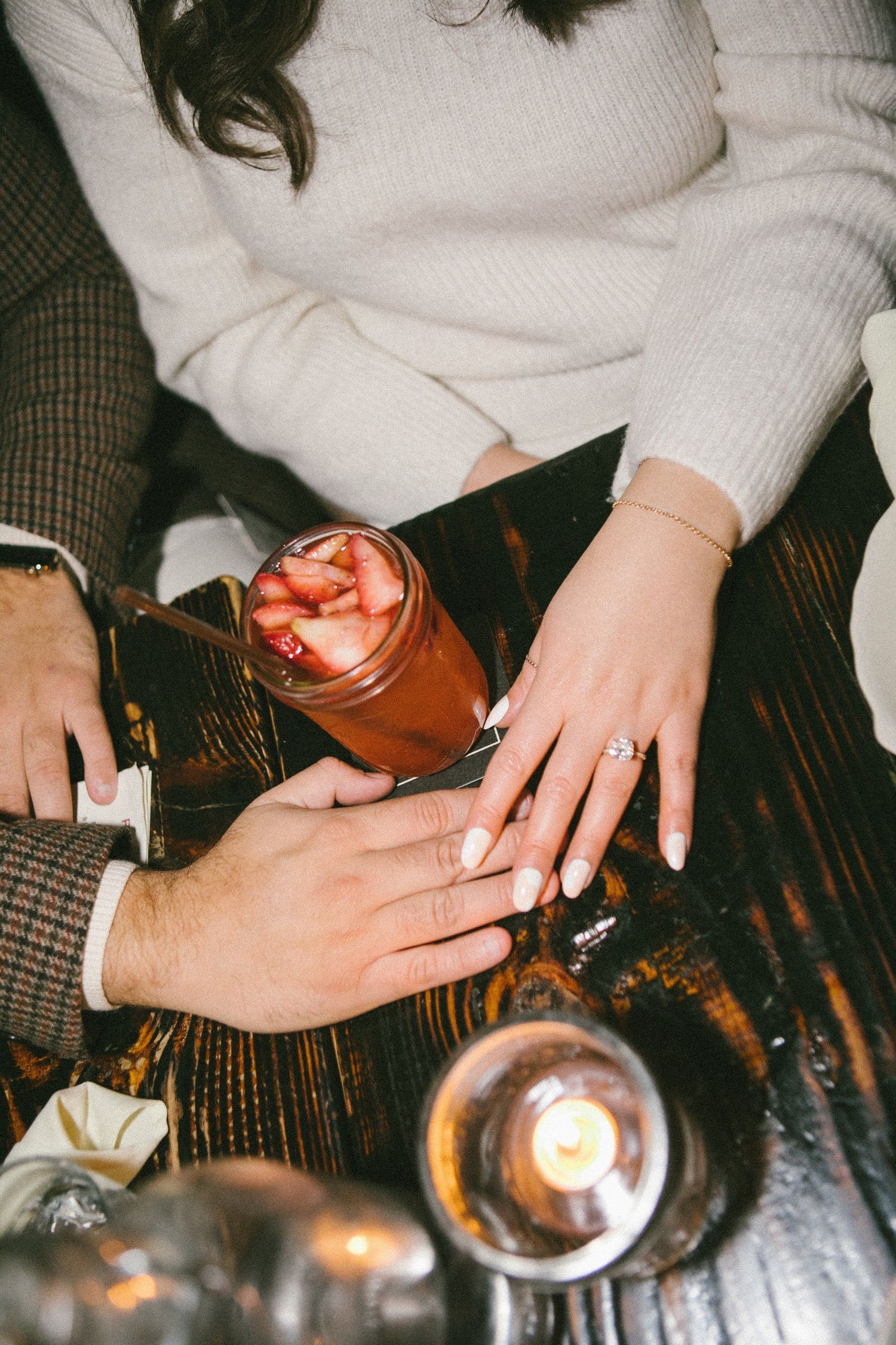 Hands with manicured nails holding drinks on a dark wooden surface with candlelight.