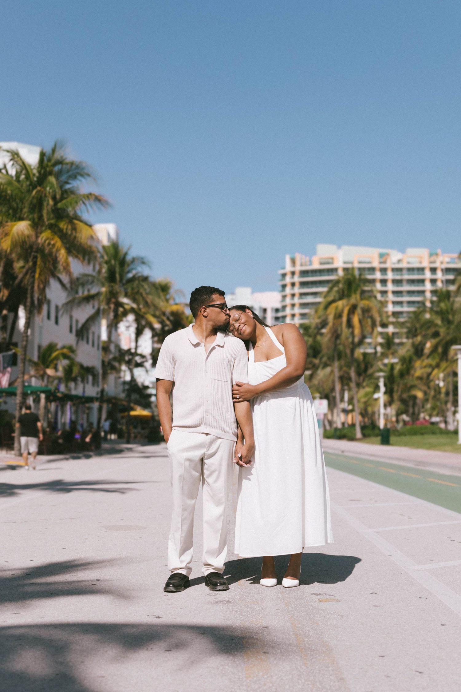 Couple in white outfits walking along palm-lined street in tropical location.