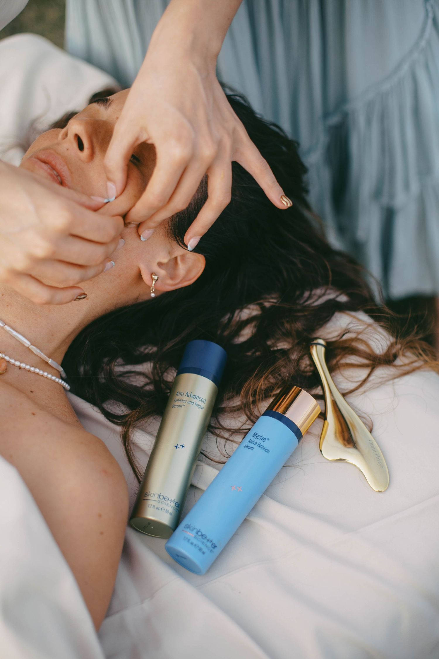 Beauty products and skincare items arranged on white bedding with hands reaching into frame.