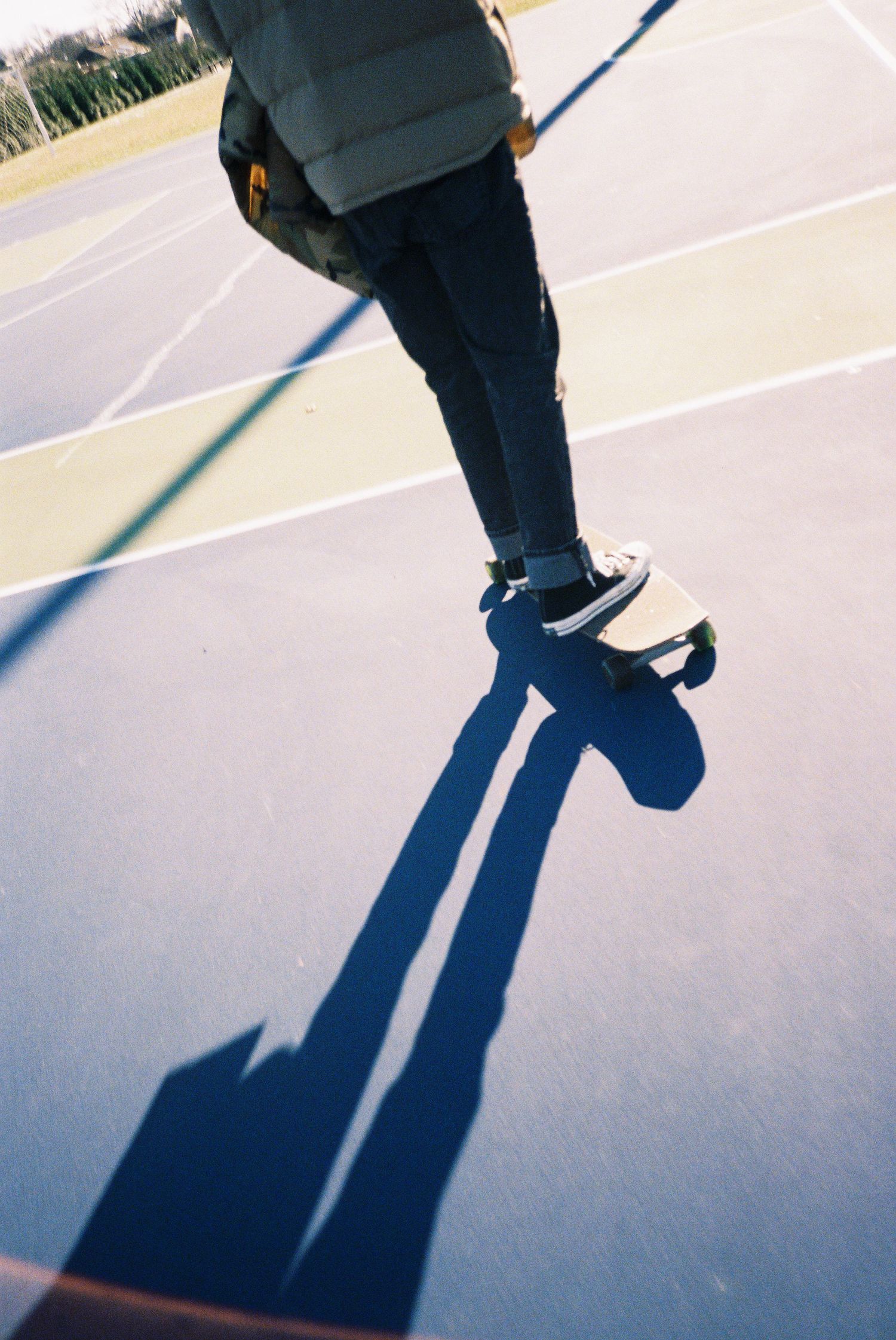 Person skateboarding on a court casting a long shadow on the pavement in sunny outdoor conditions.