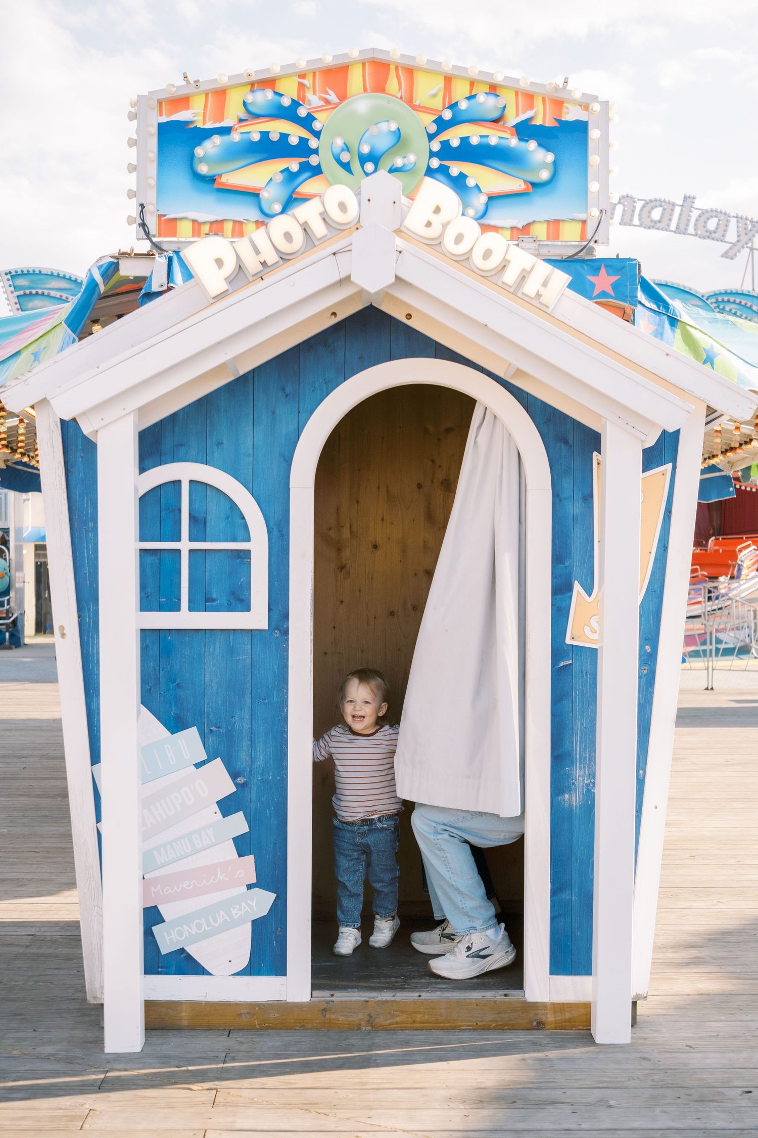 A toddler stands inside a blue and white photo booth at an amusement park on a sunny day.