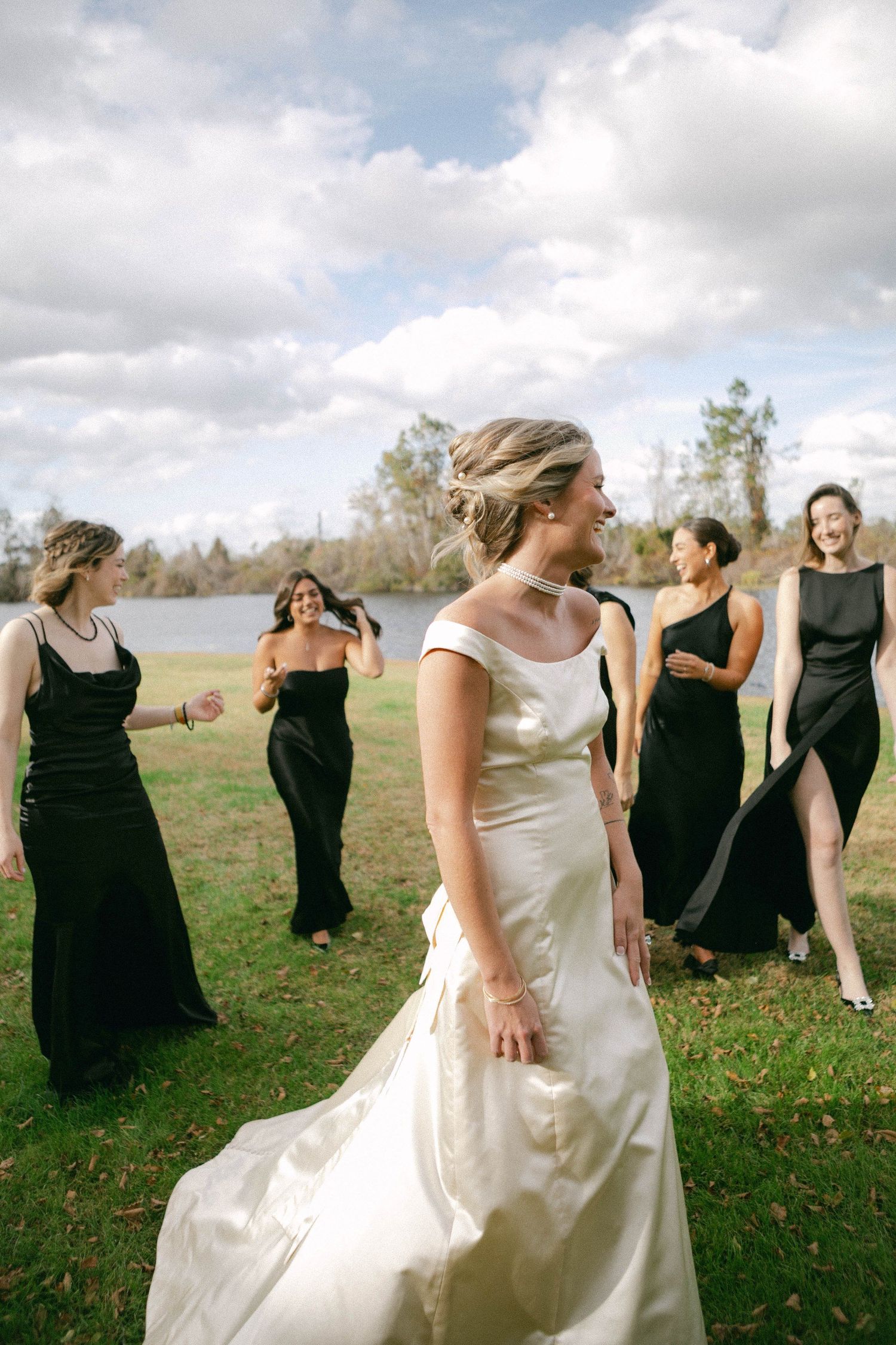 Wedding party in black dresses and white gown dancing outdoors under cloudy skies.