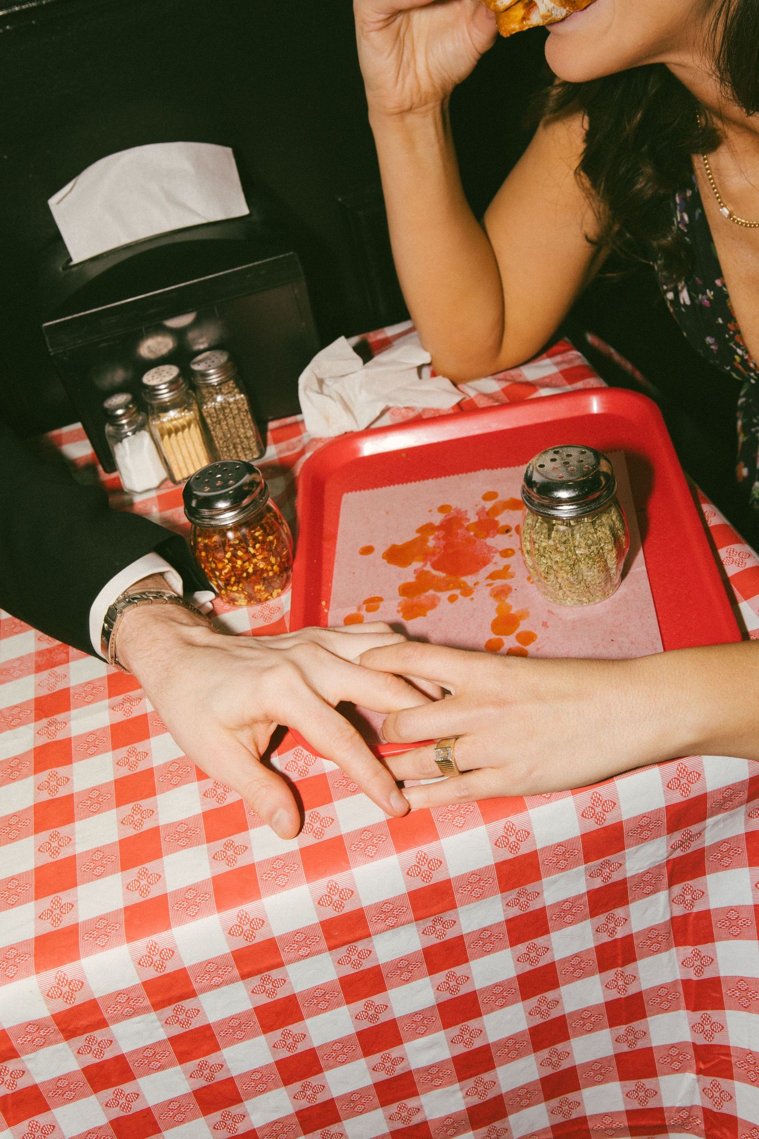 Couple holding hands on red gingham tablecloth at diner, with salt shakers and red tray visible.