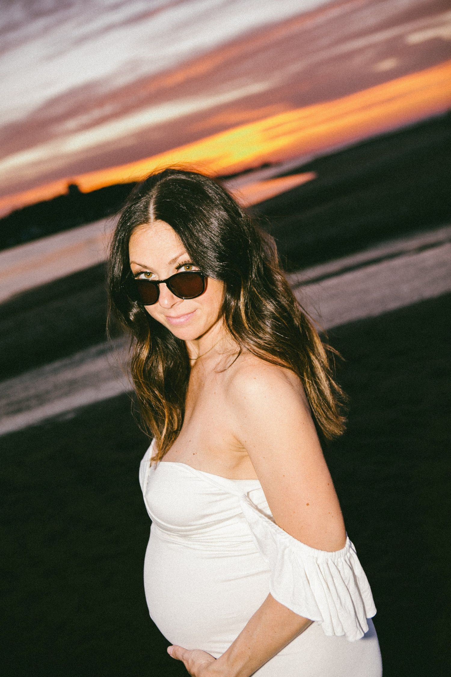 Woman in white dress posing against dramatic sunset sky at the beach.