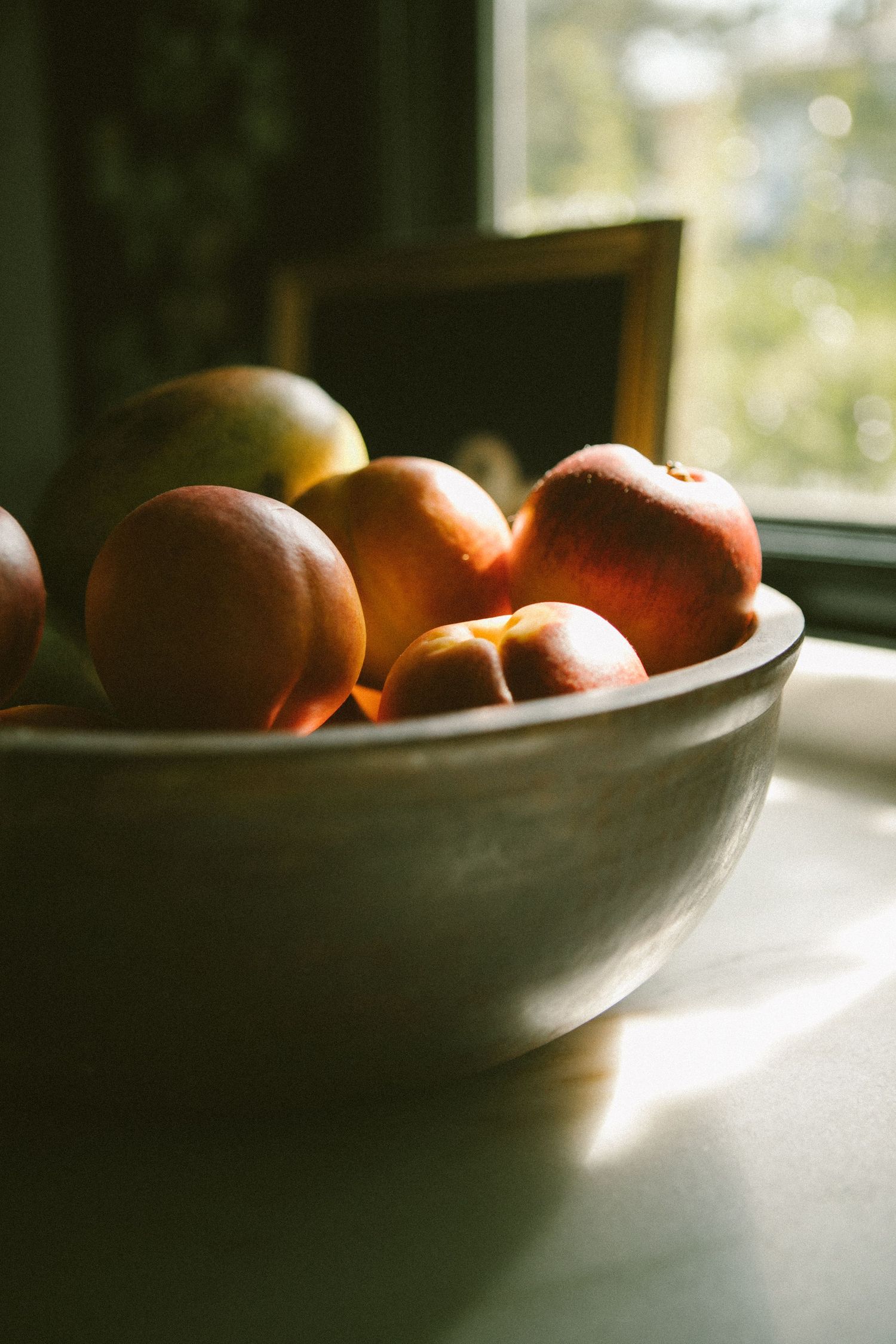 Soft natural light illuminates fresh oranges in a dark ceramic bowl by a window, creating a moody still life composition.