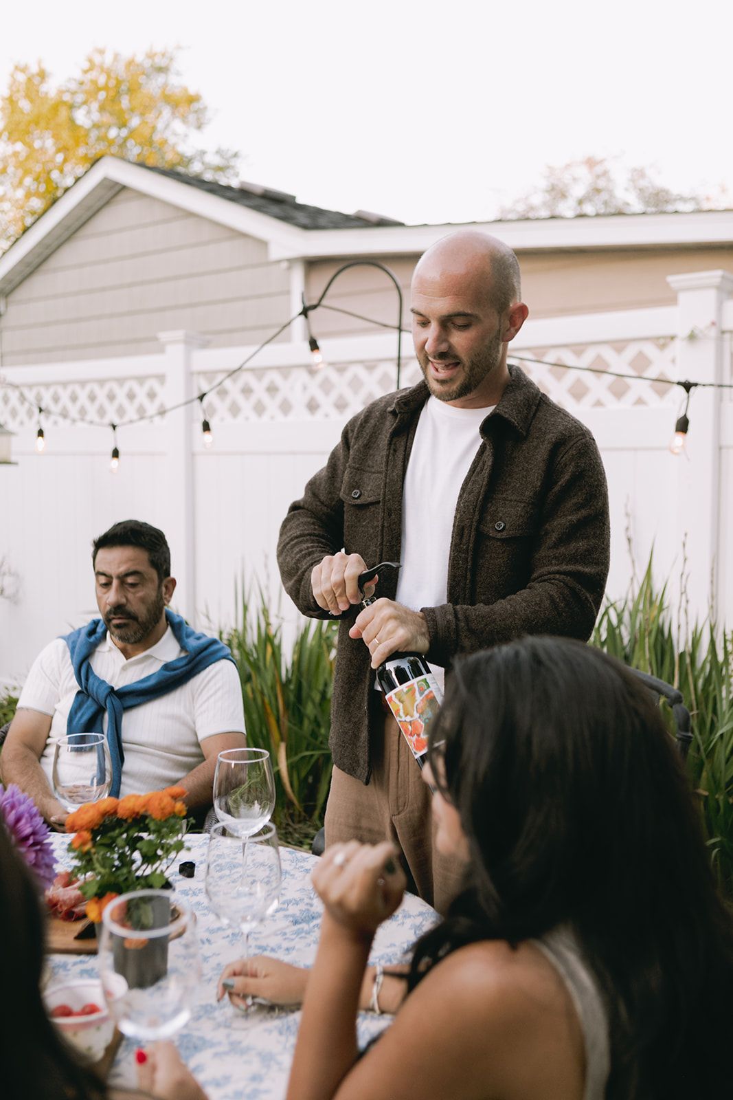 Friends gather around an outdoor table for a casual backyard celebration with string lights overhead.