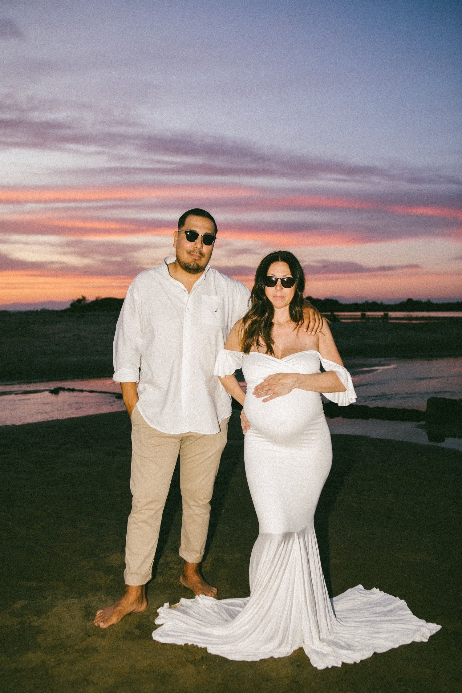 Couple in white outfits posing on beach during dramatic sunset.