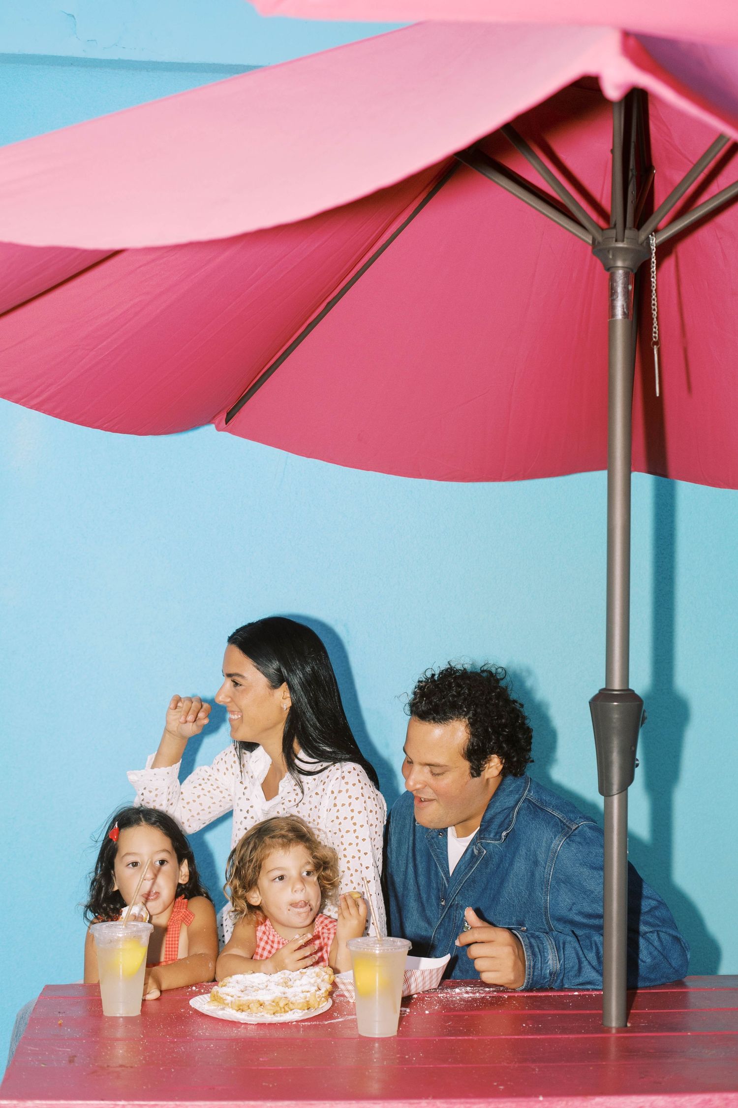 Family enjoying drinks and snacks under a pink umbrella against a bright blue wall.