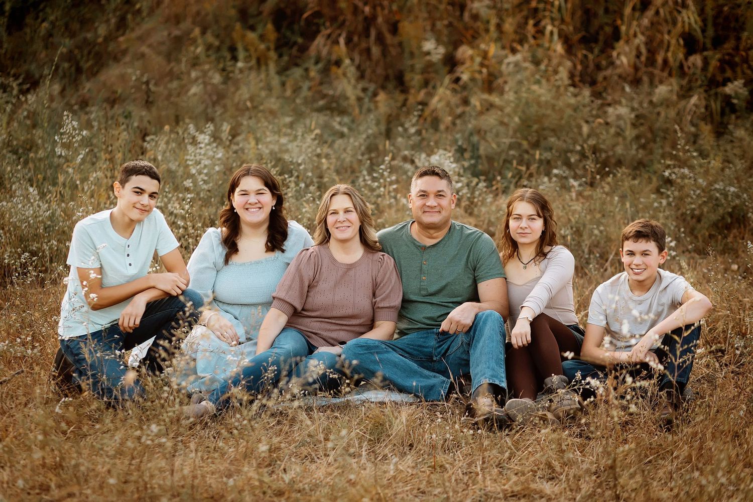 Family of six sitting together in a golden autumn field, smiling warmly for an outdoor portrait photo.