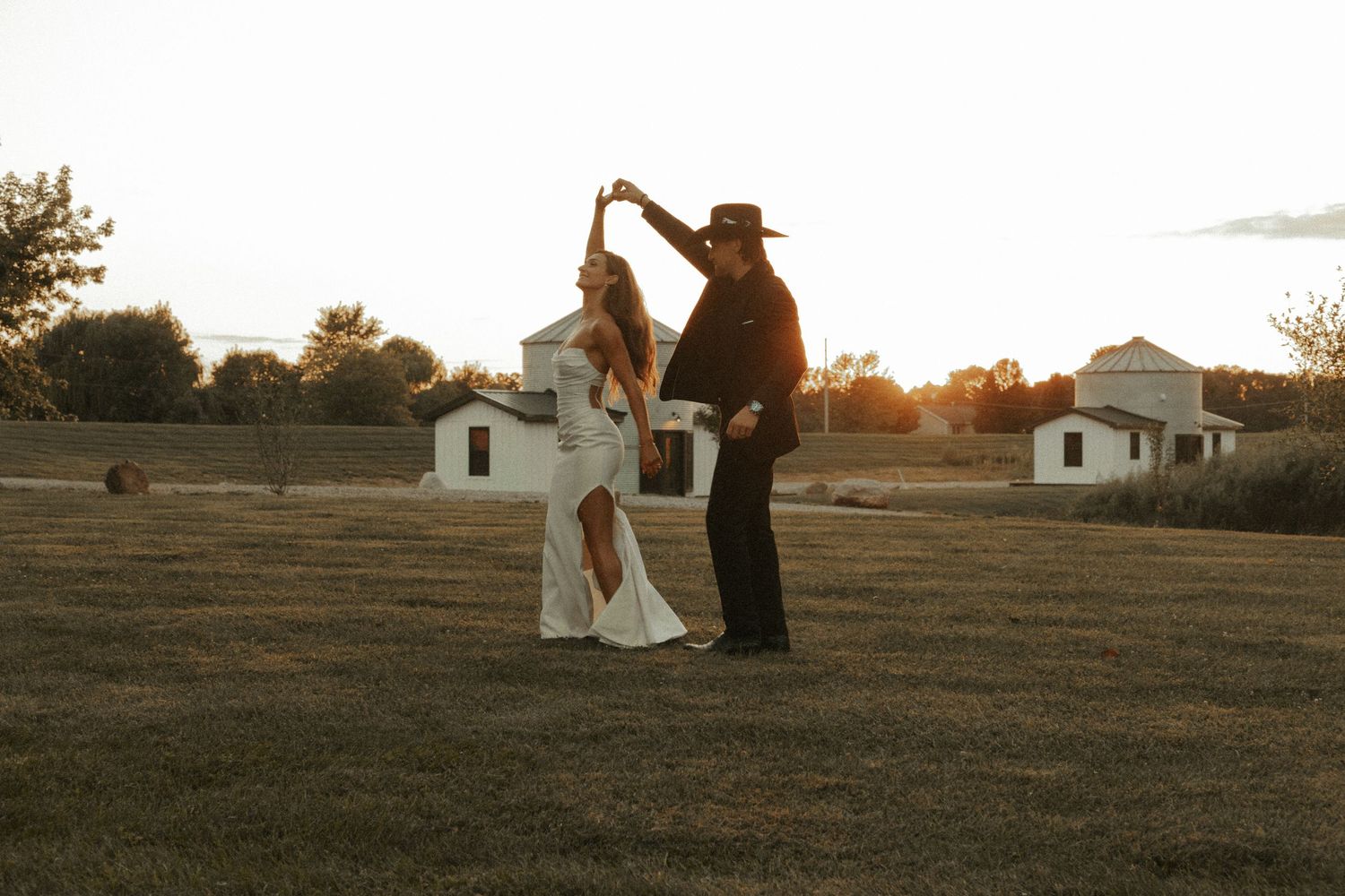 A couple dances together in an open field at sunset with small white buildings visible in the background.