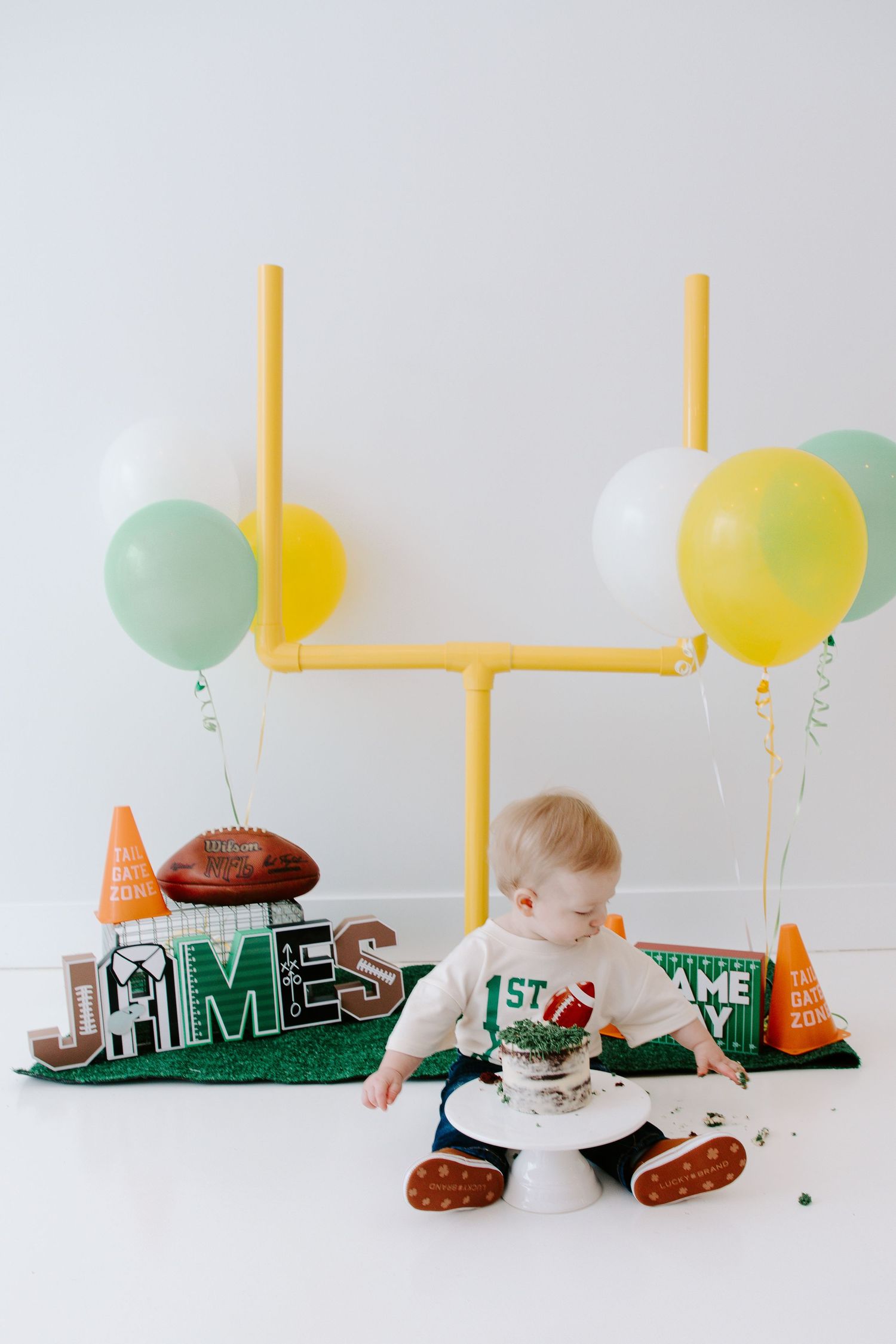 Baby in white sitting by football-themed decorations with yellow and mint green balloons.