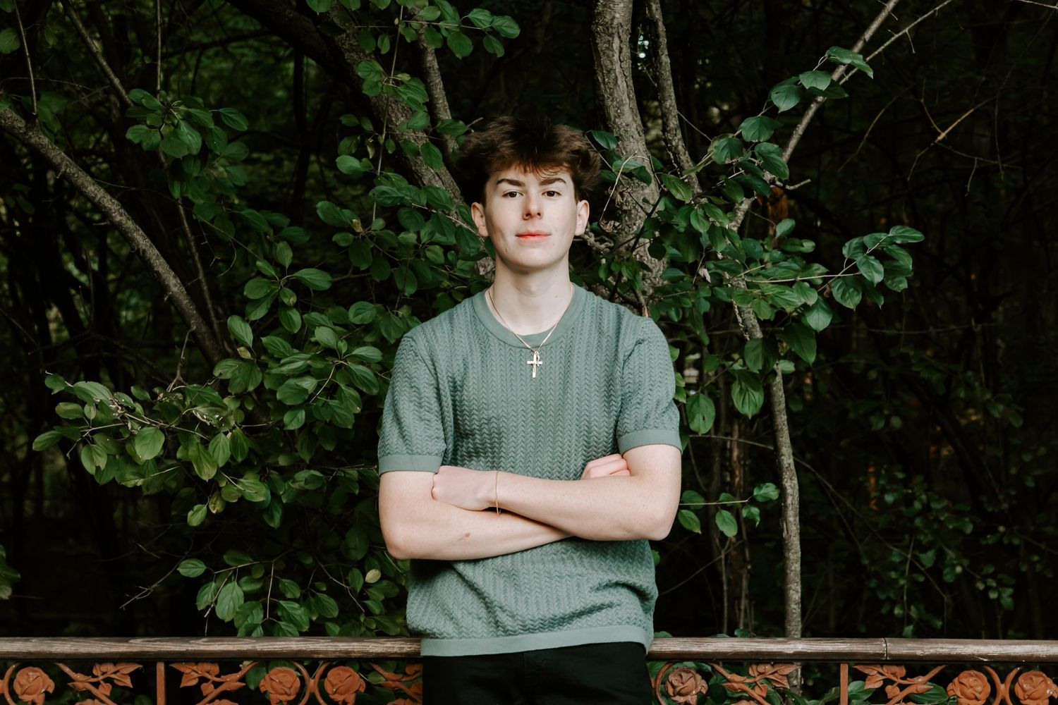 Teen in grey sweater standing against wooded backdrop with arms crossed.