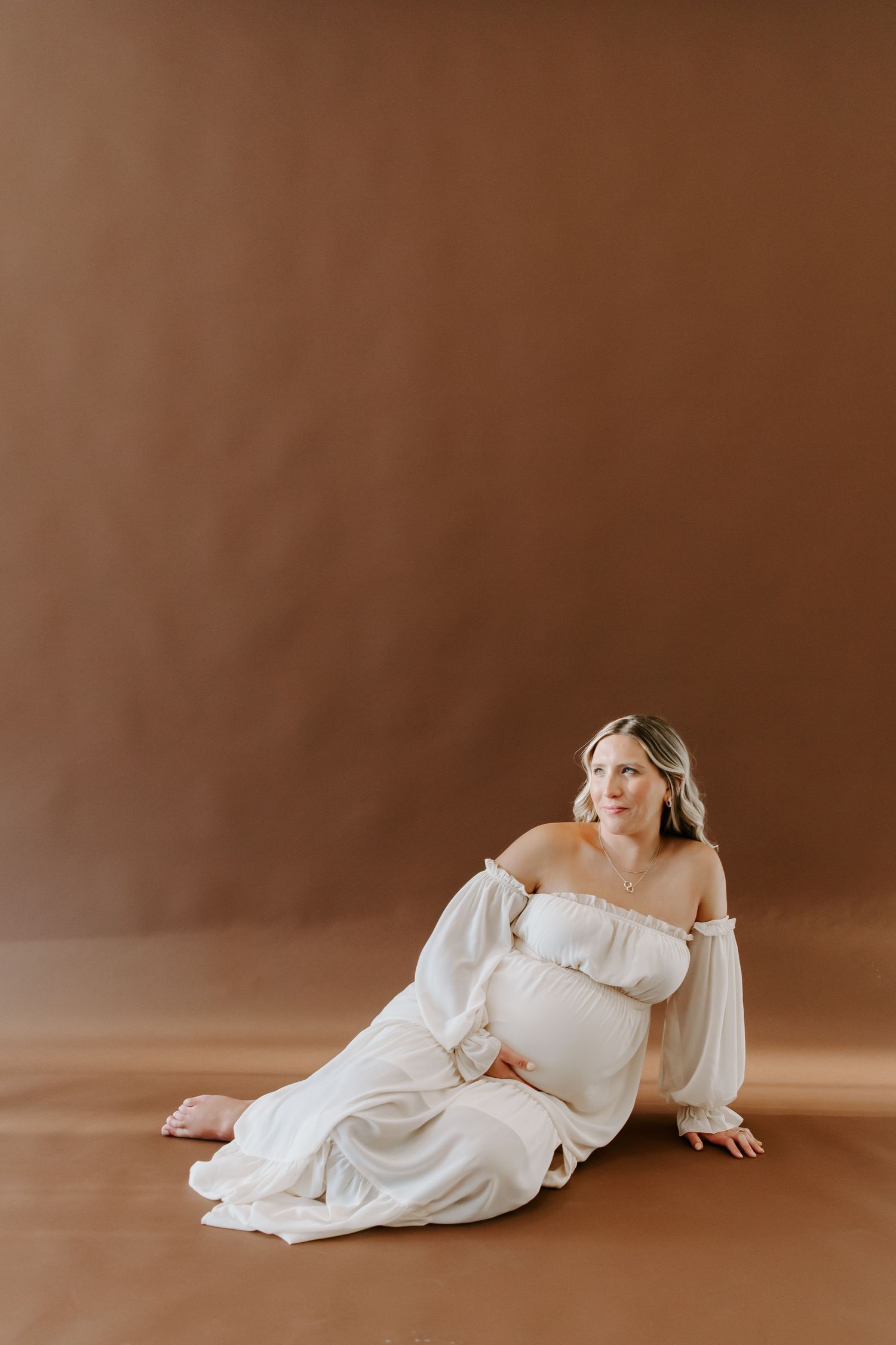 Artistic maternity portrait in white off-shoulder dress against brown background.